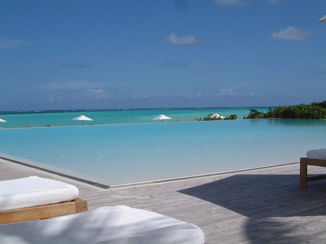 A serene infinity pool overlooks a turquoise ocean under a clear blue sky. White sun loungers with towels and white umbrellas line the wooden deck, creating a tranquil, tropical atmosphere.