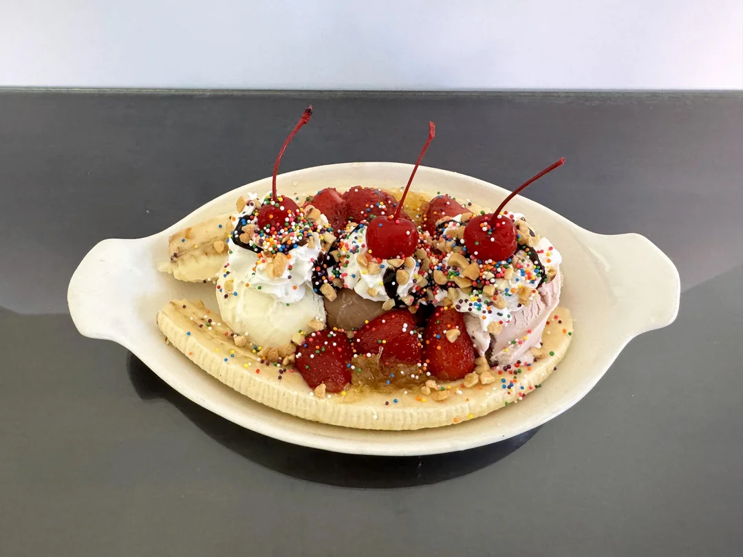 A banana split dessert with three scoops of ice cream, whipped cream, cherries, strawberries, chopped nuts, and colorful sprinkles in a white dish on a dark surface.