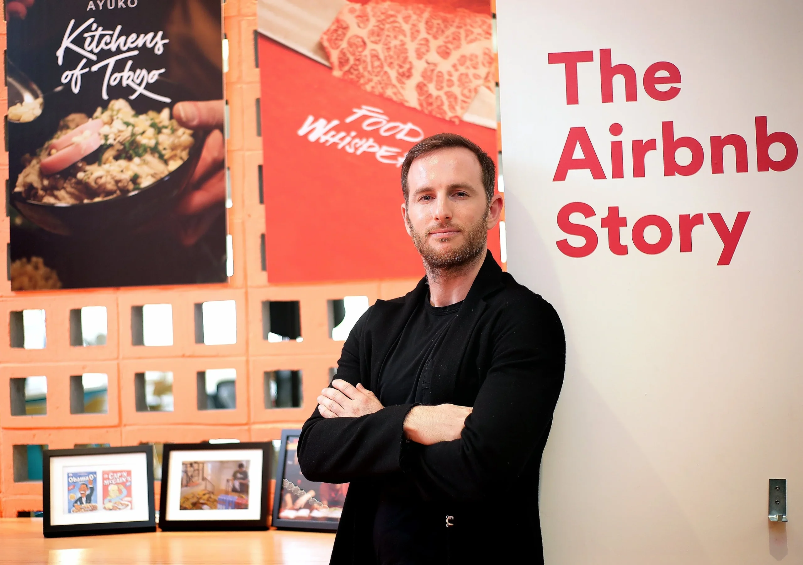 A man in a black jacket stands with arms crossed in front of a wall that says "The Airbnb Story," with framed photos and food posters in the background.