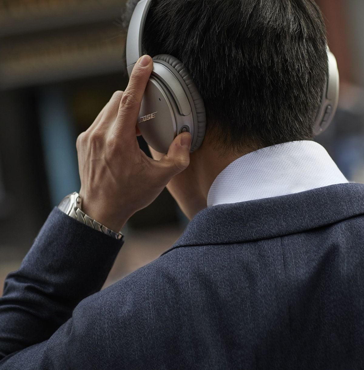 A person in a navy suit and white shirt is seen from behind, wearing over-ear Bose headphones and a metal wristwatch, with one hand touching the headphones.
