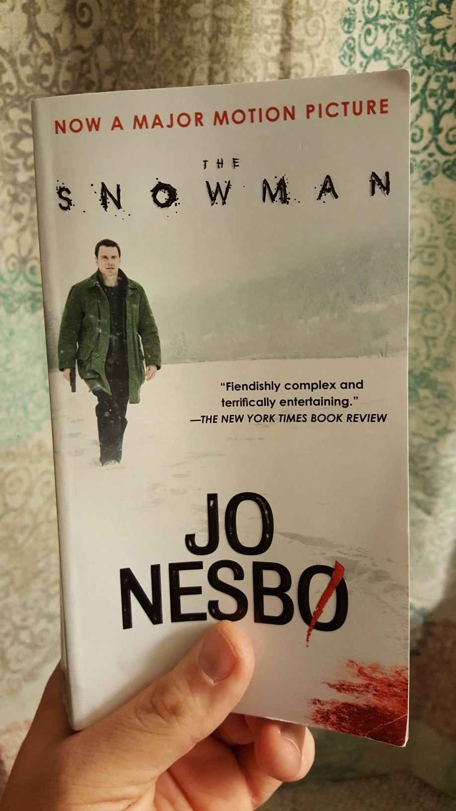 A hand holds a copy of "The Snowman" by Jo Nesbø, featuring a man in a green coat walking through snow and a red bloodstain near the author’s name. The cover notes it's now a major motion picture.