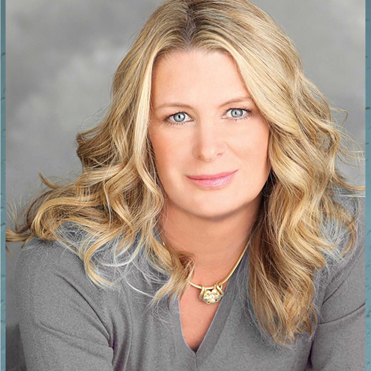 A blonde woman with wavy hair and blue eyes is wearing a gray sweater and a gold necklace, smiling softly at the camera against a gray, blurred background.