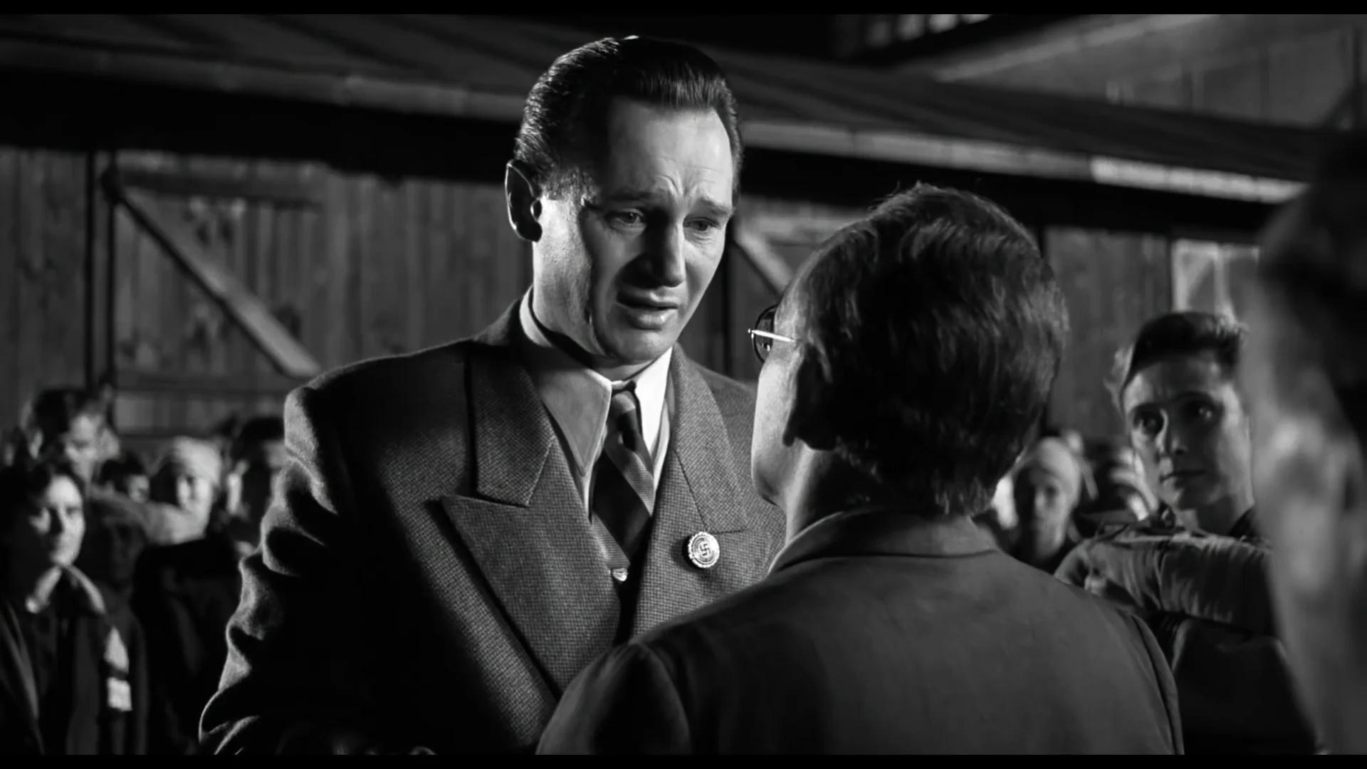 A man in a suit speaks emotionally to another person in glasses in a black-and-white scene. Other people stand in the background, watching the interaction. The setting appears to be outdoors near a wooden building.
