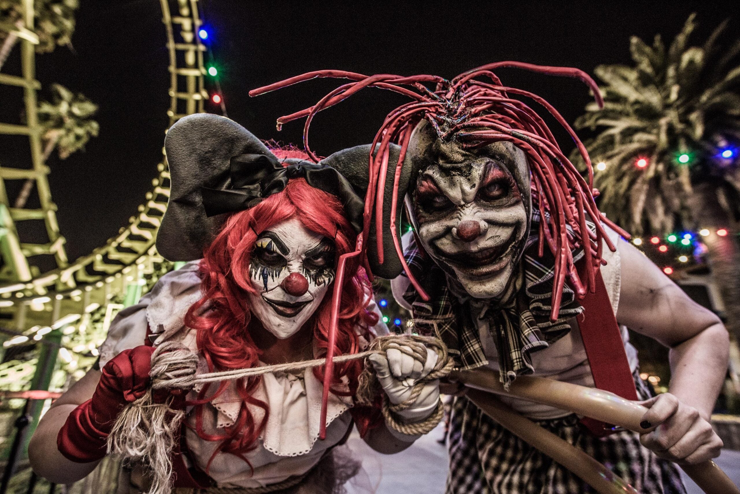 Two people dressed as creepy clowns with smeared face paint, red noses, and wild red hair pose menacingly at night in front of a roller coaster and colorful string lights. Palm trees are visible in the background.