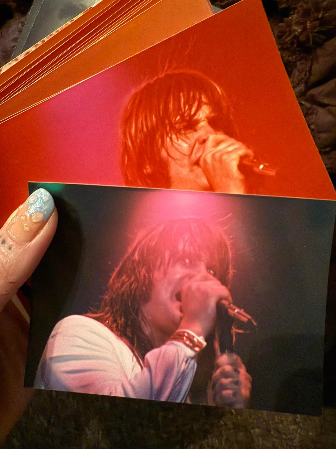 A hand with painted nails holds two glossy photos of a long-haired singer performing on stage, both images showing the singer with a microphone and intense stage lighting.