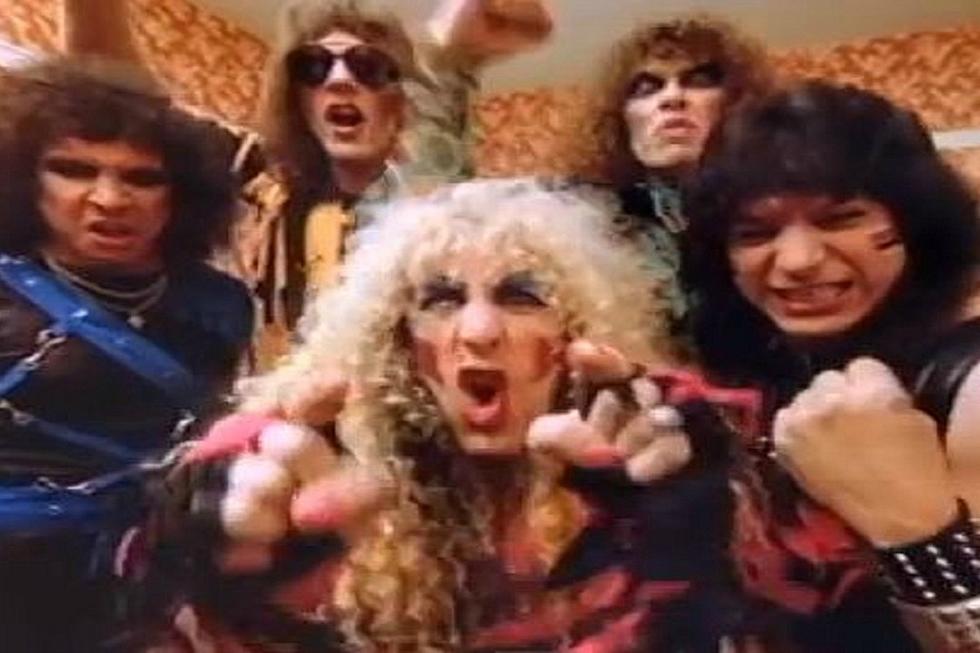 Five people with big, teased hair and dramatic makeup, wearing colorful, edgy clothes, make intense faces and gestures toward the camera in a lively, patterned room, evoking an 80s rock band style.