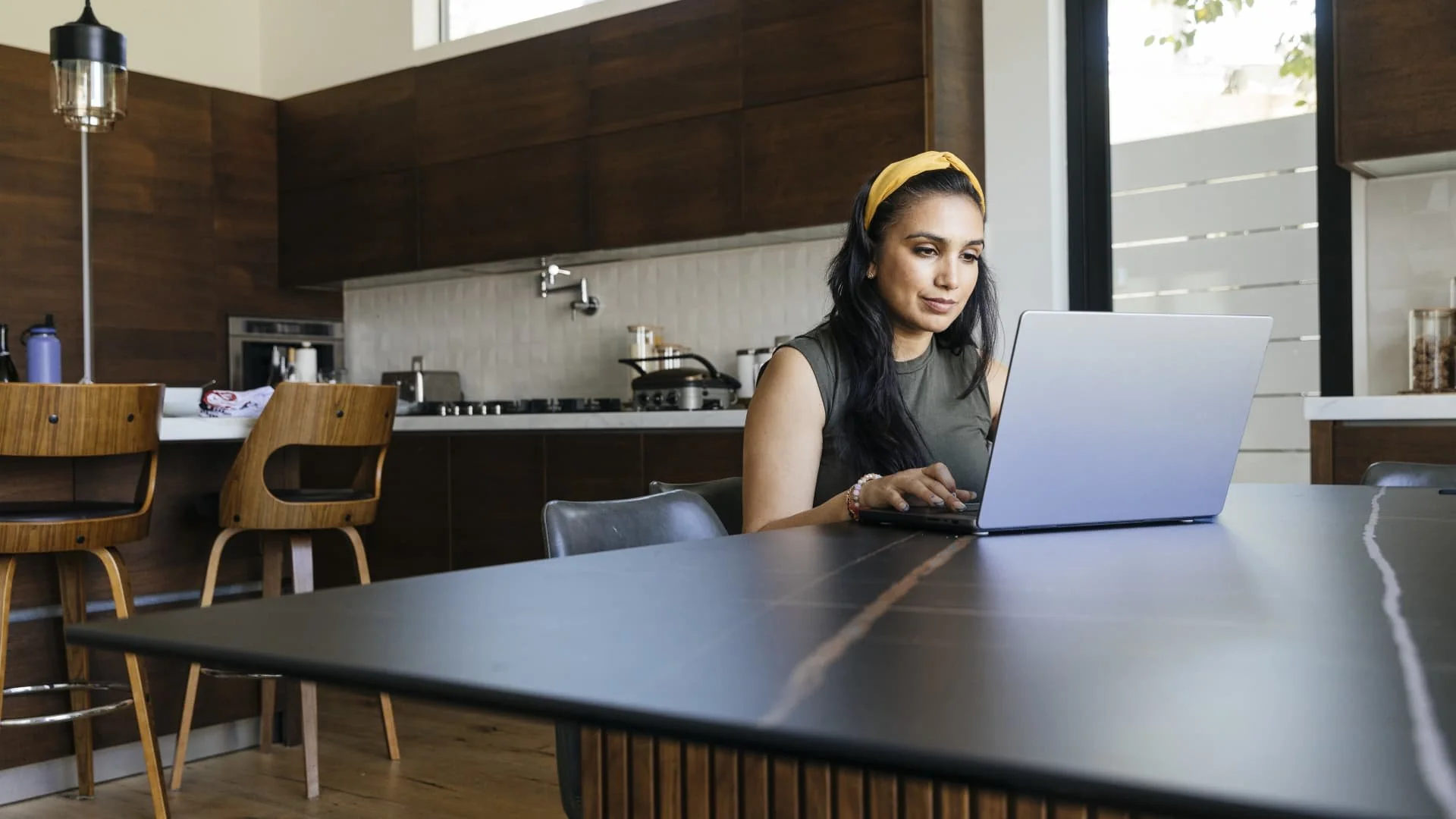 A woman with long dark hair and a yellow headband works on a laptop at a table in a modern kitchen with wooden cabinets and bar stools in the background.