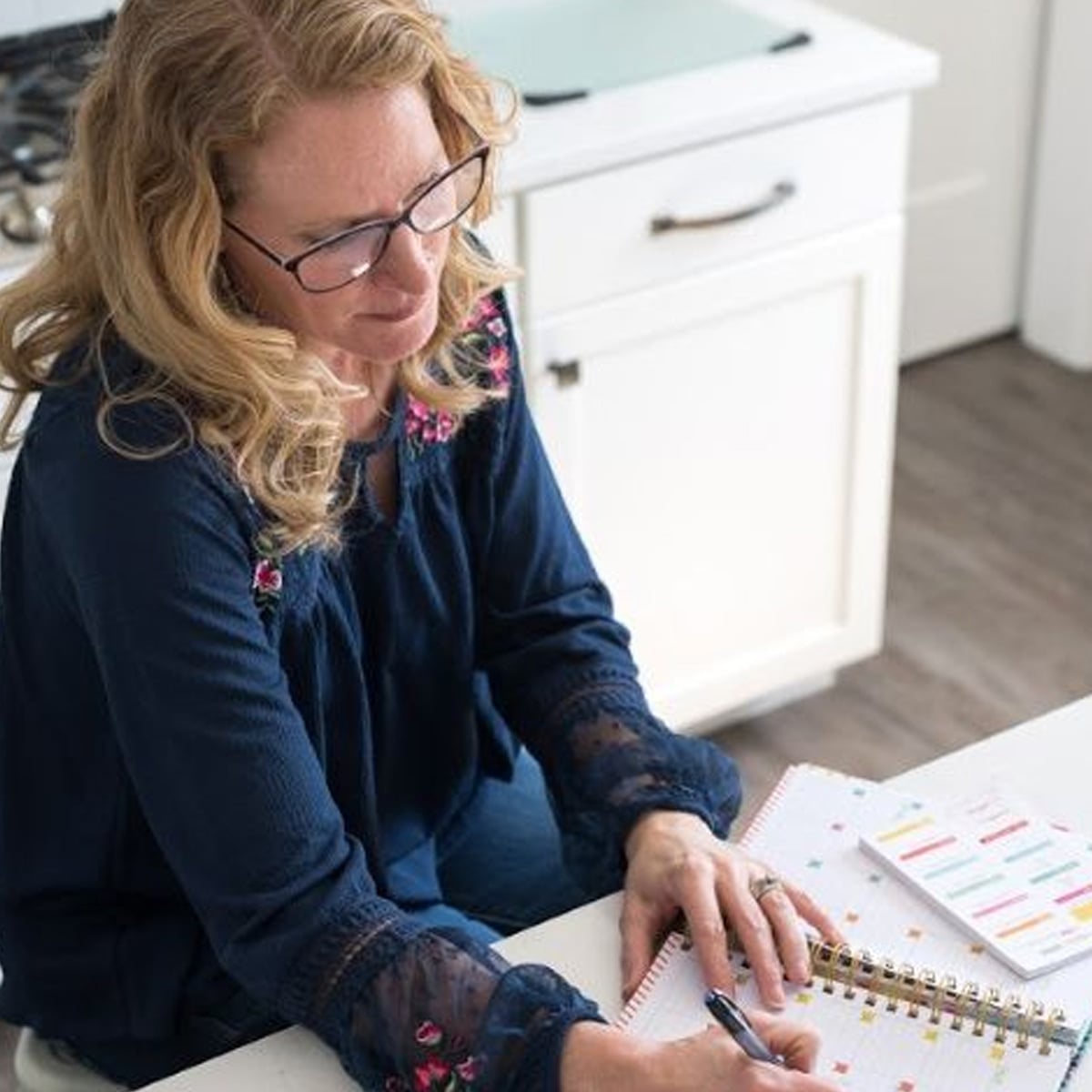 A woman with blonde hair and glasses sits at a kitchen counter, writing in a planner or notebook. She wears a blue top with embroidered details and appears focused on her work.