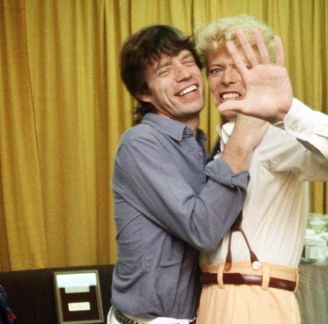 Two smiling men stand close together in front of yellow curtains; one hugs the other, who playfully holds up his hand toward the camera as if blocking the view.