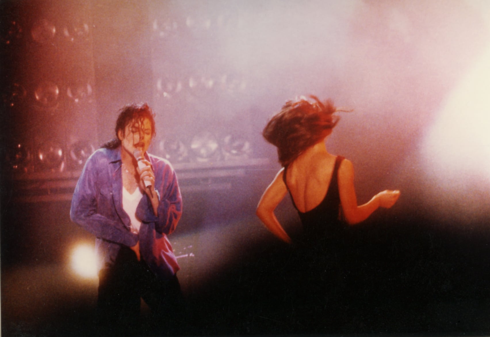 A male singer in a blue shirt performs passionately on stage, holding a microphone, while a woman in a black dress dances nearby; colored stage lights and smoke create a dramatic atmosphere.