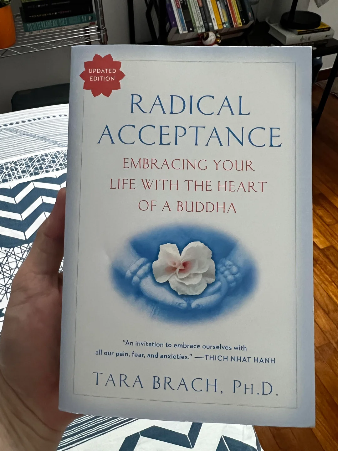 A hand holds the book "Radical Acceptance: Embracing Your Life with the Heart of a Buddha" by Tara Brach, Ph.D., with a white cover, a flower image, and a quote from Thich Nhat Hanh.