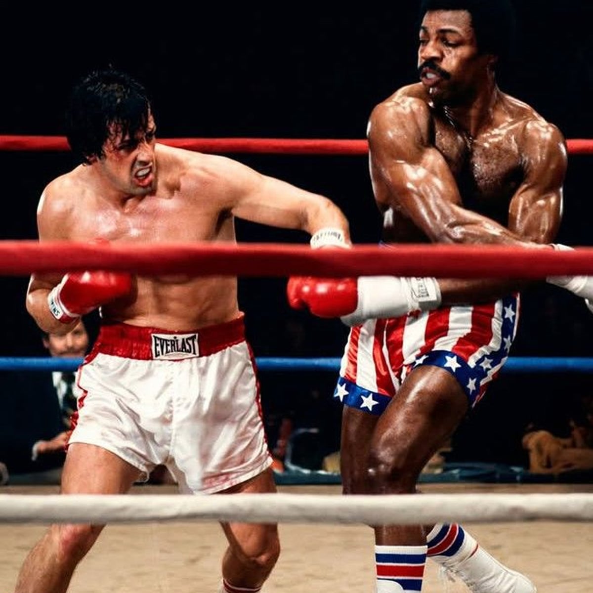 Two male boxers are fighting in a boxing ring. One, wearing white trunks, throws a punch at his opponent, who wears red, white, and blue star-spangled shorts. Both look intense and athletic under bright lights.