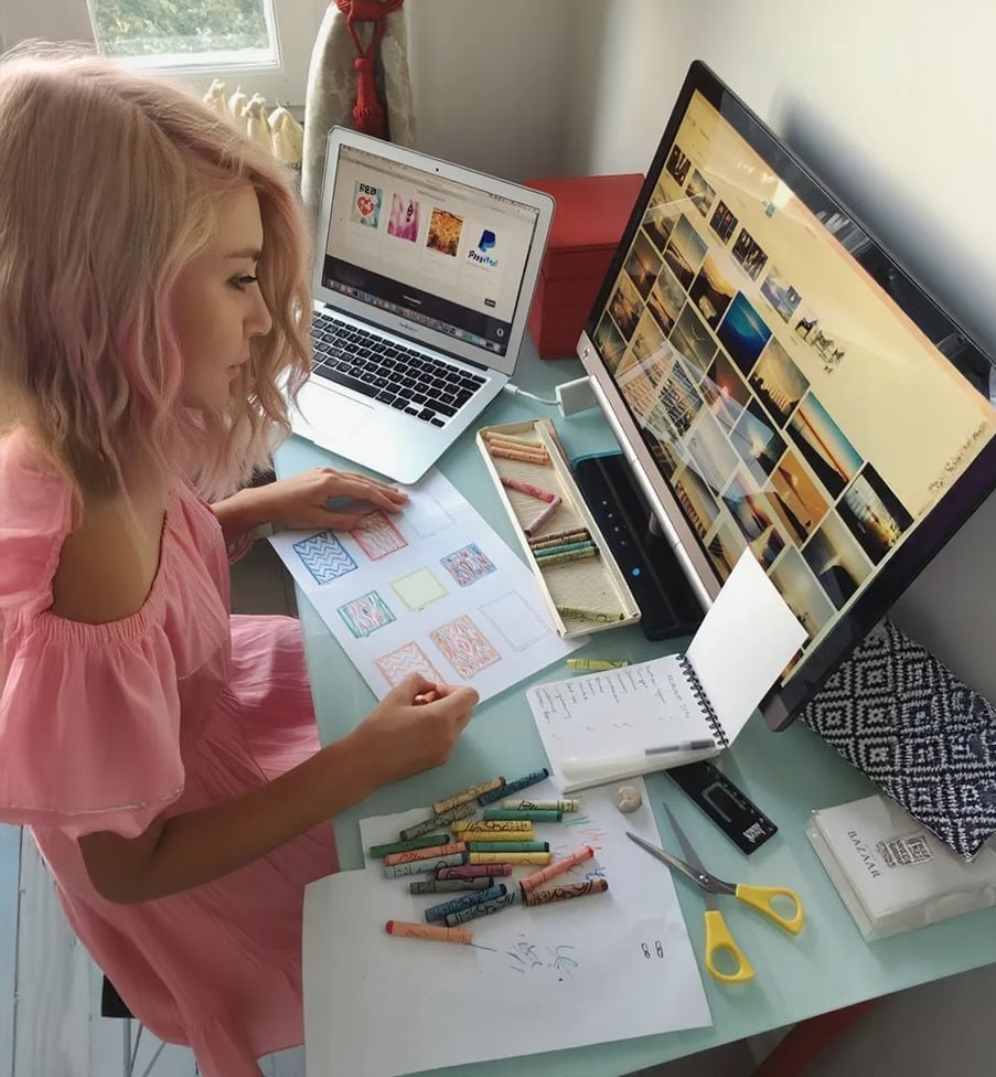 A woman with pink hair in a pink dress sits at a desk with crayons, drawings, and notes, working between a laptop and a large monitor displaying image thumbnails.