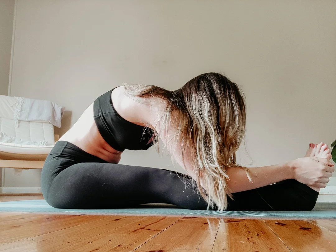 A woman in black athletic wear sits on a yoga mat indoors, stretching forward to touch her toes with both hands, her head resting near her knees. The floor is wooden, and a chair is visible in the background.