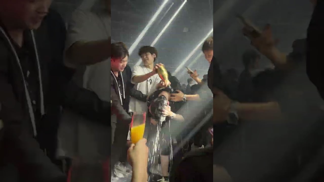 A group of people at a party pour drinks over someone’s head while others cheer and hold up glasses. Bright lights create a festive atmosphere.