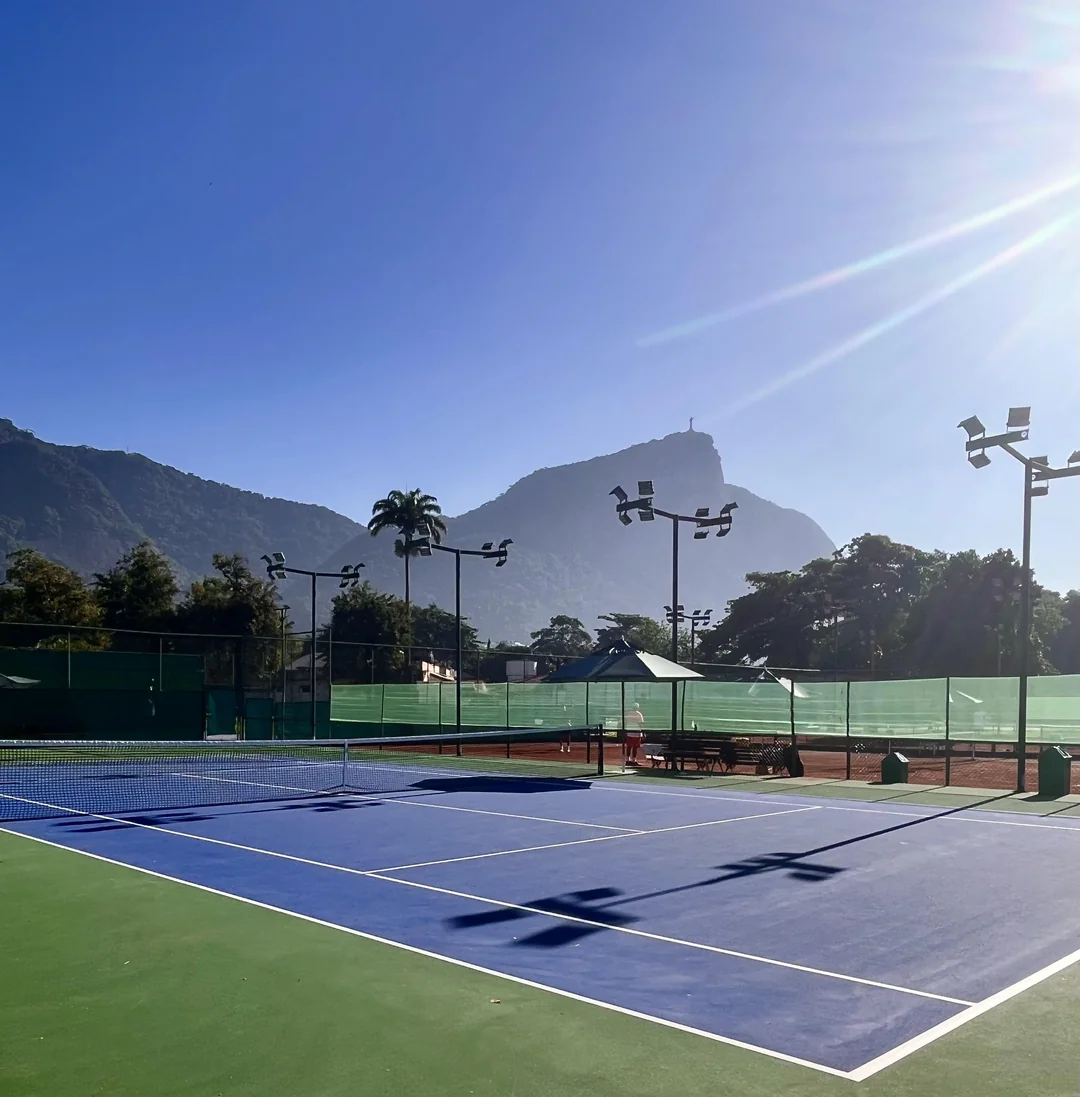 A sunlit outdoor tennis court with blue and green surfaces, surrounded by fences and lamp posts, with mountains and palm trees in the background under a clear blue sky.