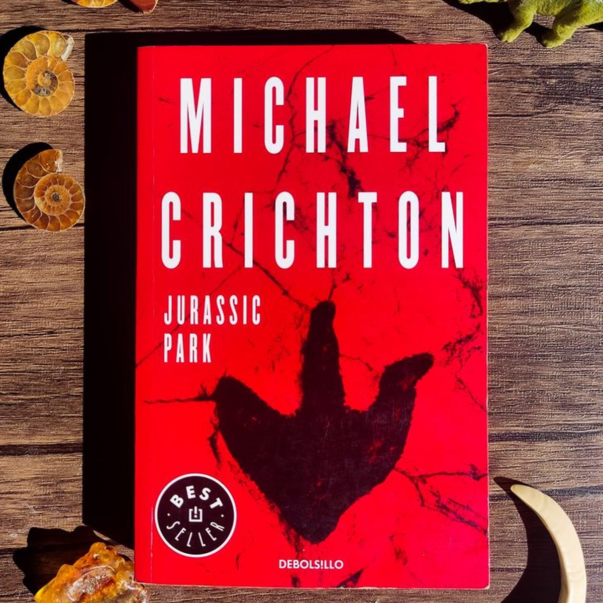 A red cover edition of "Jurassic Park" by Michael Crichton lies on a wooden surface, featuring a large black dinosaur footprint. Fossils and rocks are placed around the book.