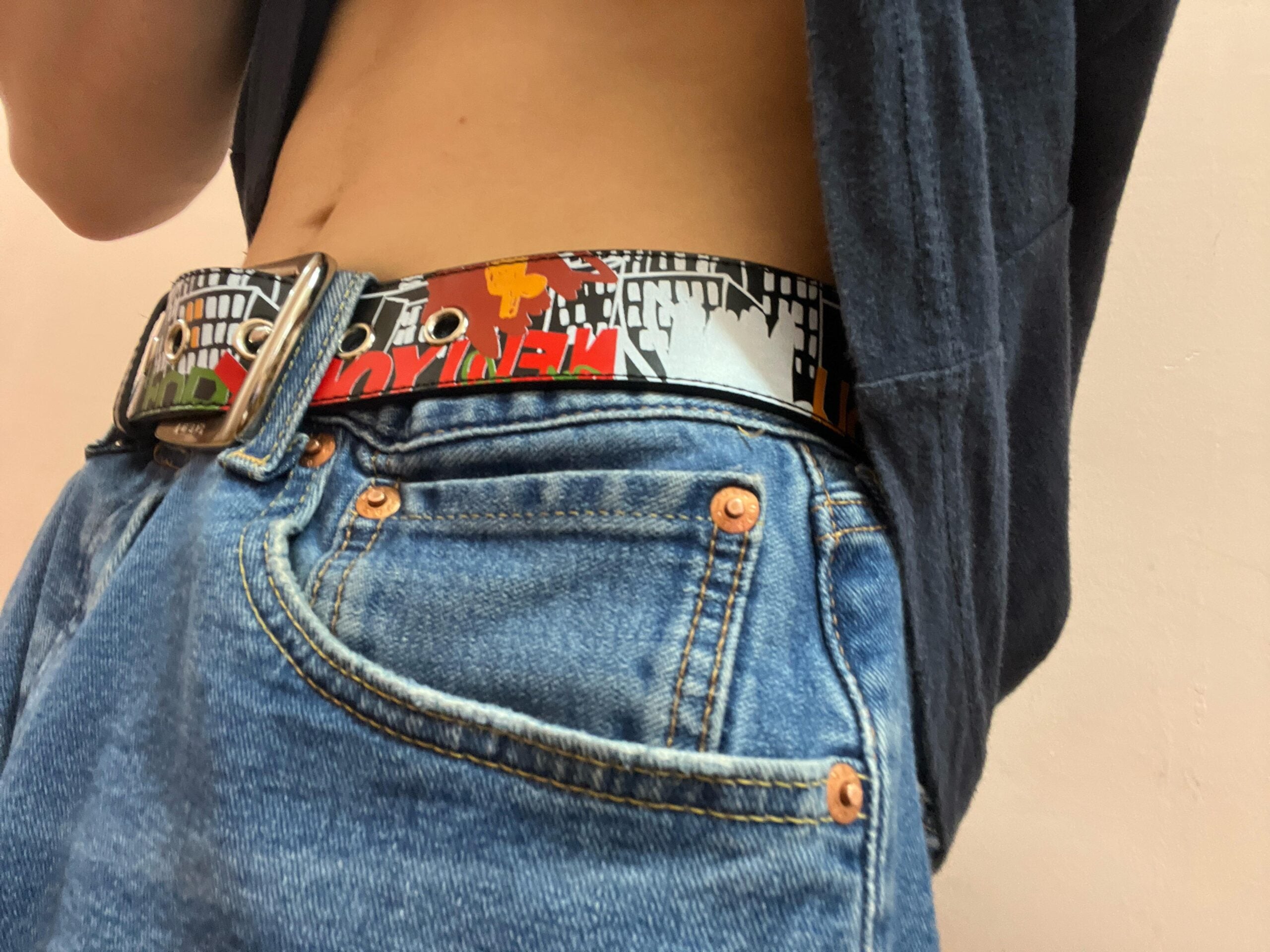 A person wearing blue jeans and a colorful, graphic-patterned belt stands with their shirt slightly lifted, showing part of their waist against a plain, light-colored background.