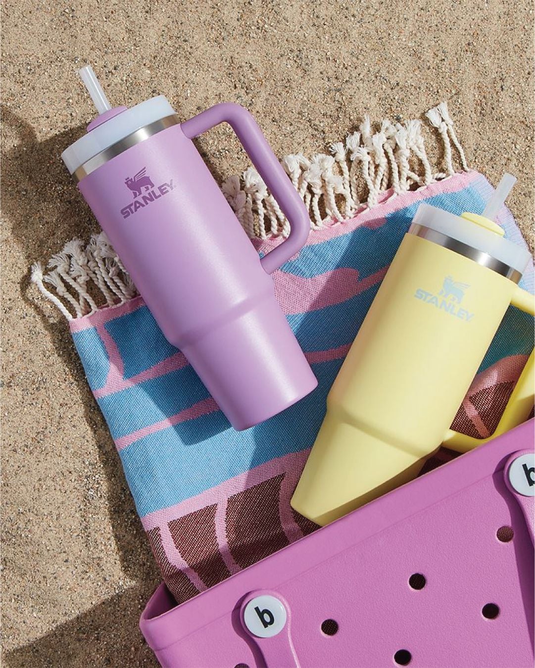 Two Stanley tumblers, one lavender and one yellow, with straws, rest on a pink and blue fringed towel on sand next to a pink Crocs-style tote bag.
