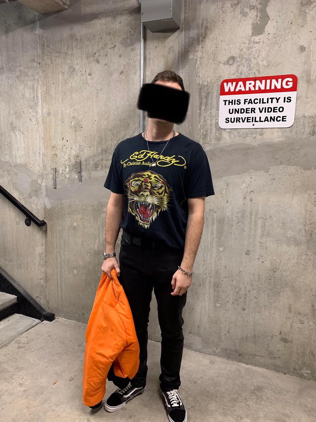 A person stands indoors near a concrete wall with a "WARNING: THIS FACILITY IS UNDER VIDEO SURVEILLANCE" sign. They wear a black graphic T-shirt, black pants, black sneakers, and hold an orange jacket. Their face is obscured.