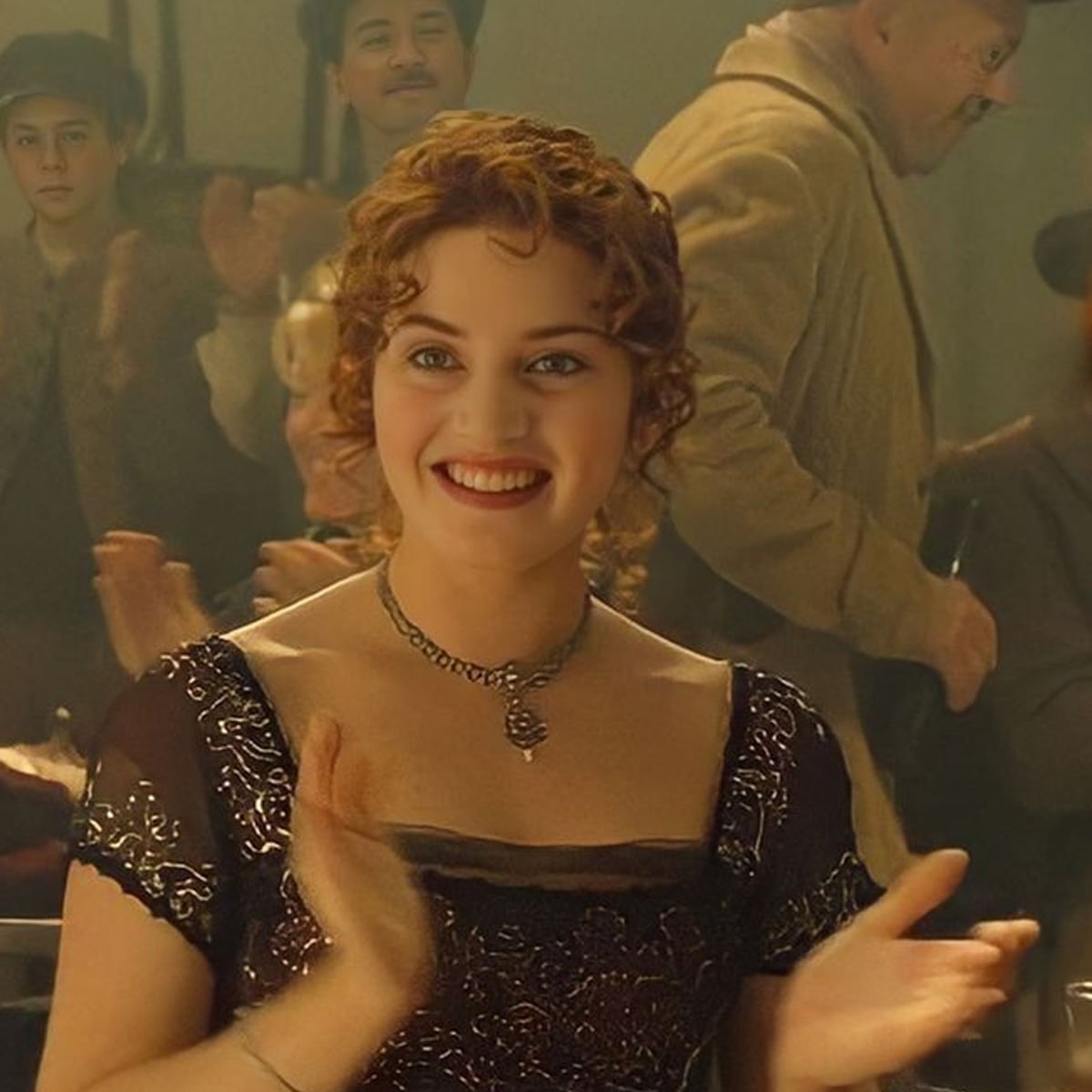 A woman with curly auburn hair, wearing a black embellished dress and necklace, smiles and claps in a warmly lit, crowded setting. Two people are visible in the blurred background.