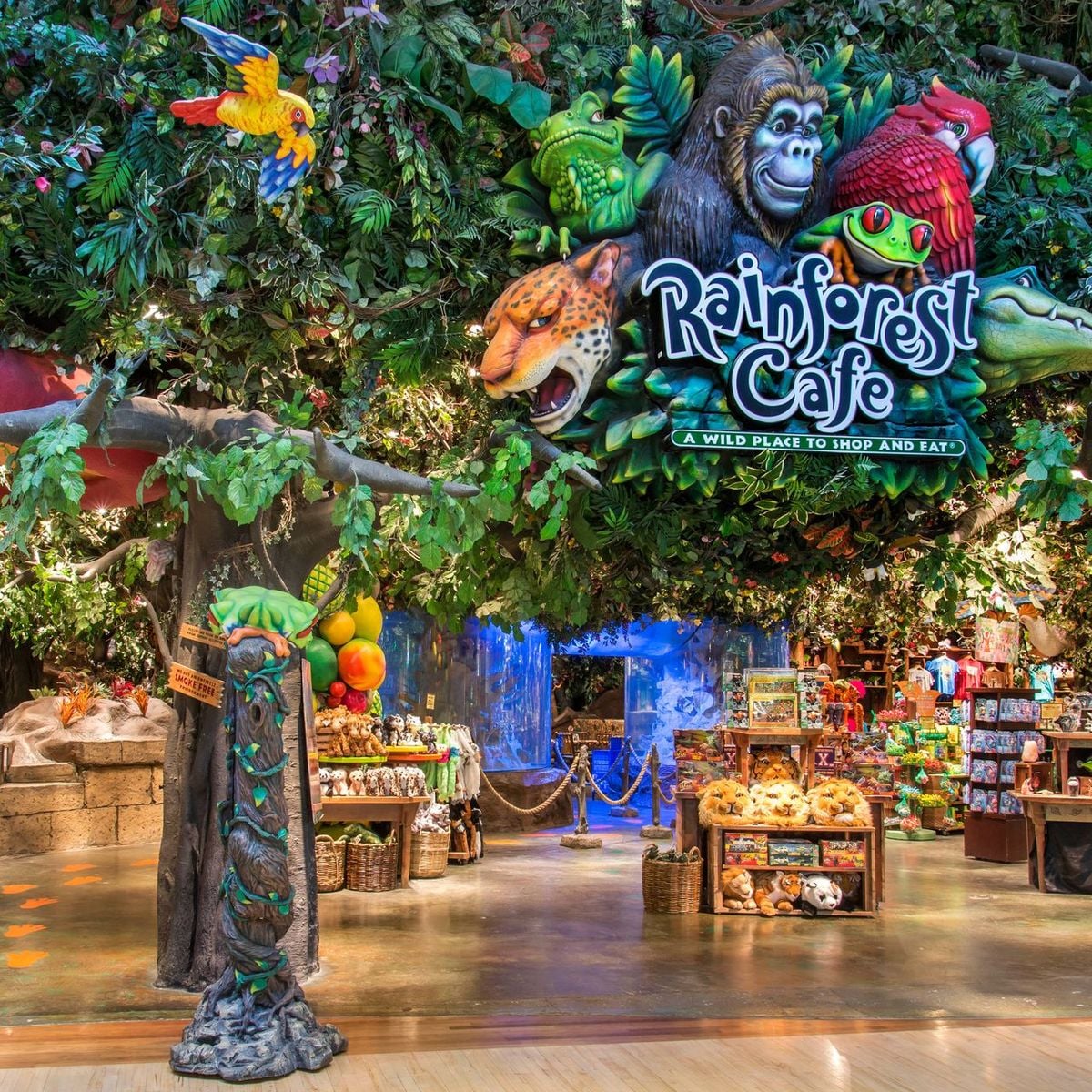 The entrance to Rainforest Cafe, decorated with lush fake greenery, animal figures like a gorilla, parrot, and jaguar, vibrant signs, and rainforest-themed merchandise displayed inside.