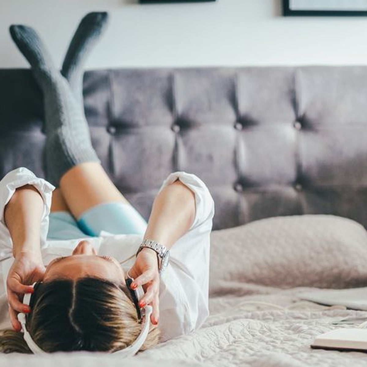 A person lies on a bed with legs up on the headboard, wearing gray knee-high socks and headphones, relaxing in a cozy bedroom setting.