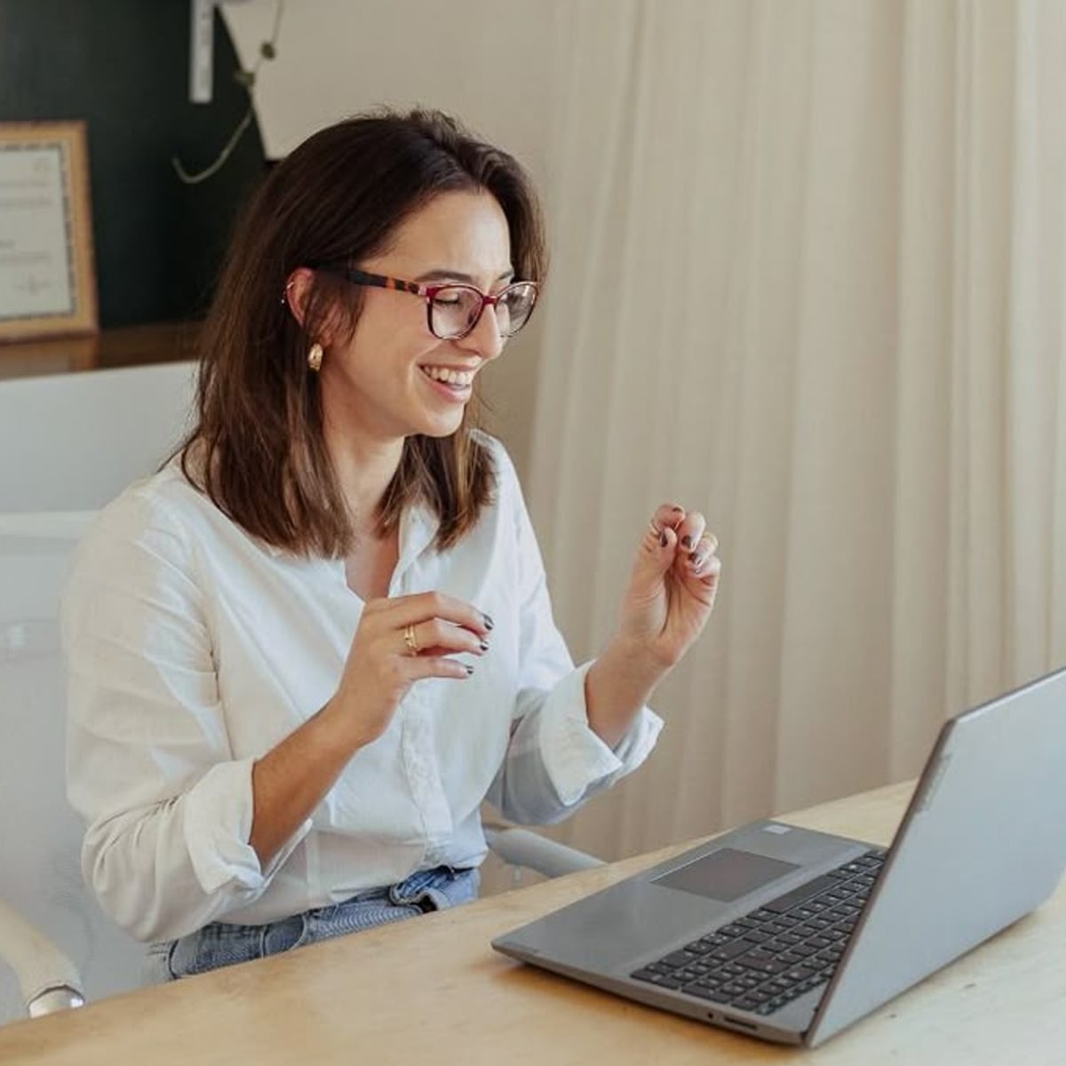 A woman wearing glasses and a white shirt smiles and gestures while sitting at a desk with an open laptop, appearing to engage in a video call in a home office setting.