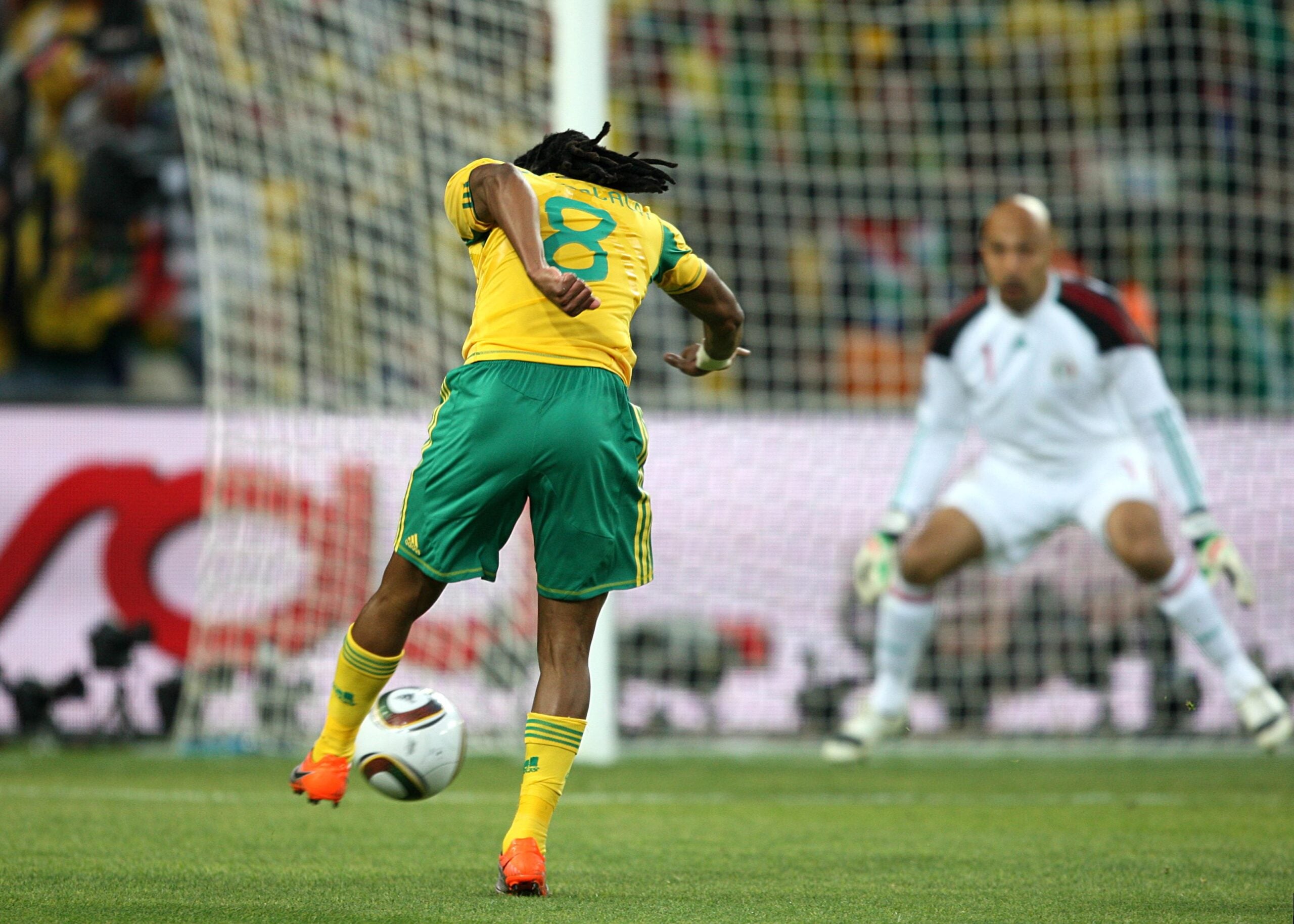 A soccer player in a yellow and green uniform prepares to kick a ball towards the goal, while a goalkeeper in white stands ready to defend in front of the net.