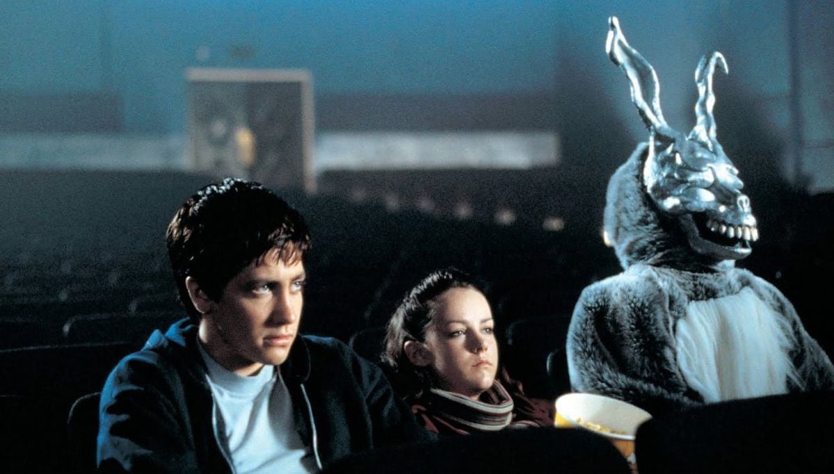 A teenage boy, a girl, and a person in a creepy rabbit costume sit in an empty, dimly lit theater. The boy looks serious, the girl is relaxed, and the rabbit figure holds a bucket of popcorn.