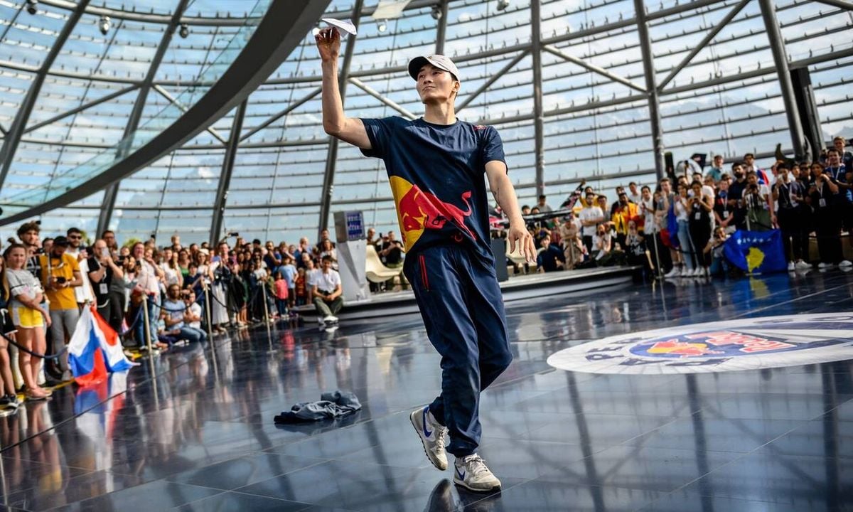 A dancer in a navy outfit and white cap performs on a shiny floor inside a glass-domed building, with a crowd watching. A Red Bull logo is visible on the dancer's shirt and on the floor.