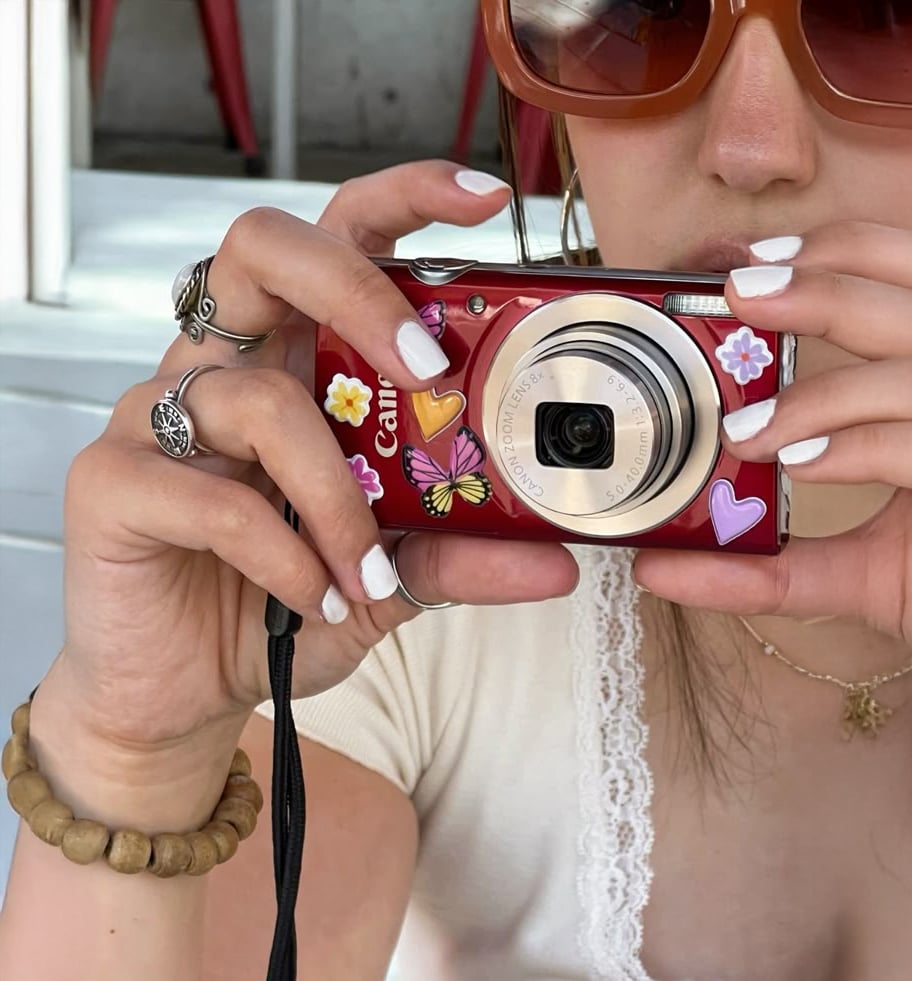 A person with white nail polish and multiple rings holds a red camera decorated with butterfly and heart stickers. They wear large sunglasses, a lace-trimmed white top, and a beaded bracelet.