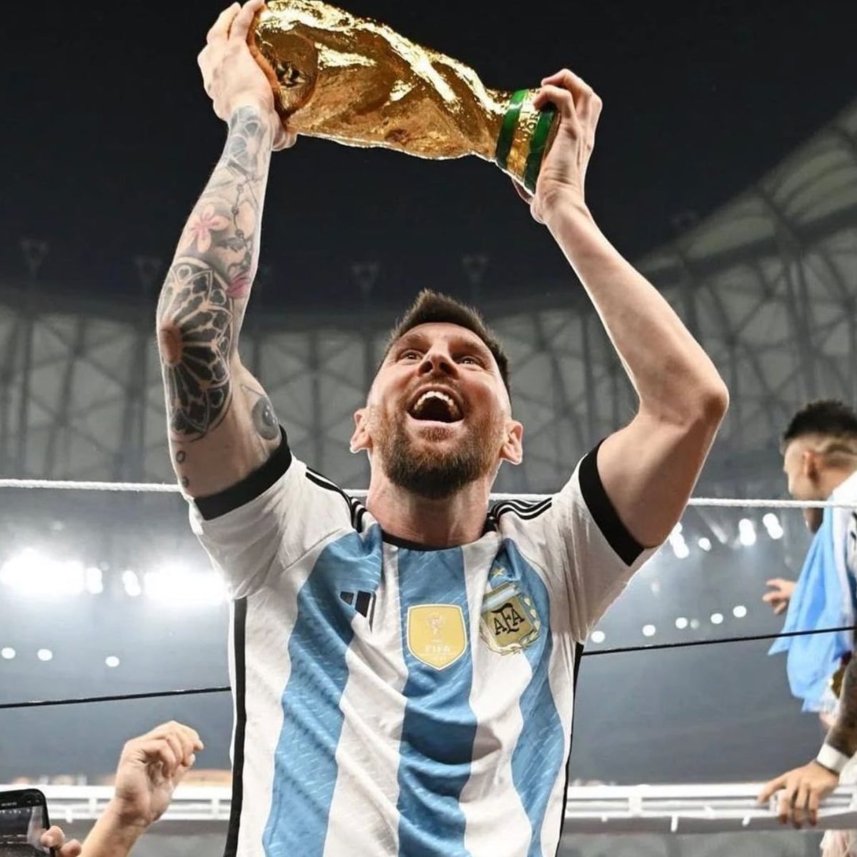 A soccer player in an Argentina jersey holds the FIFA World Cup trophy above his head, smiling joyfully in a stadium filled with lights and celebration.