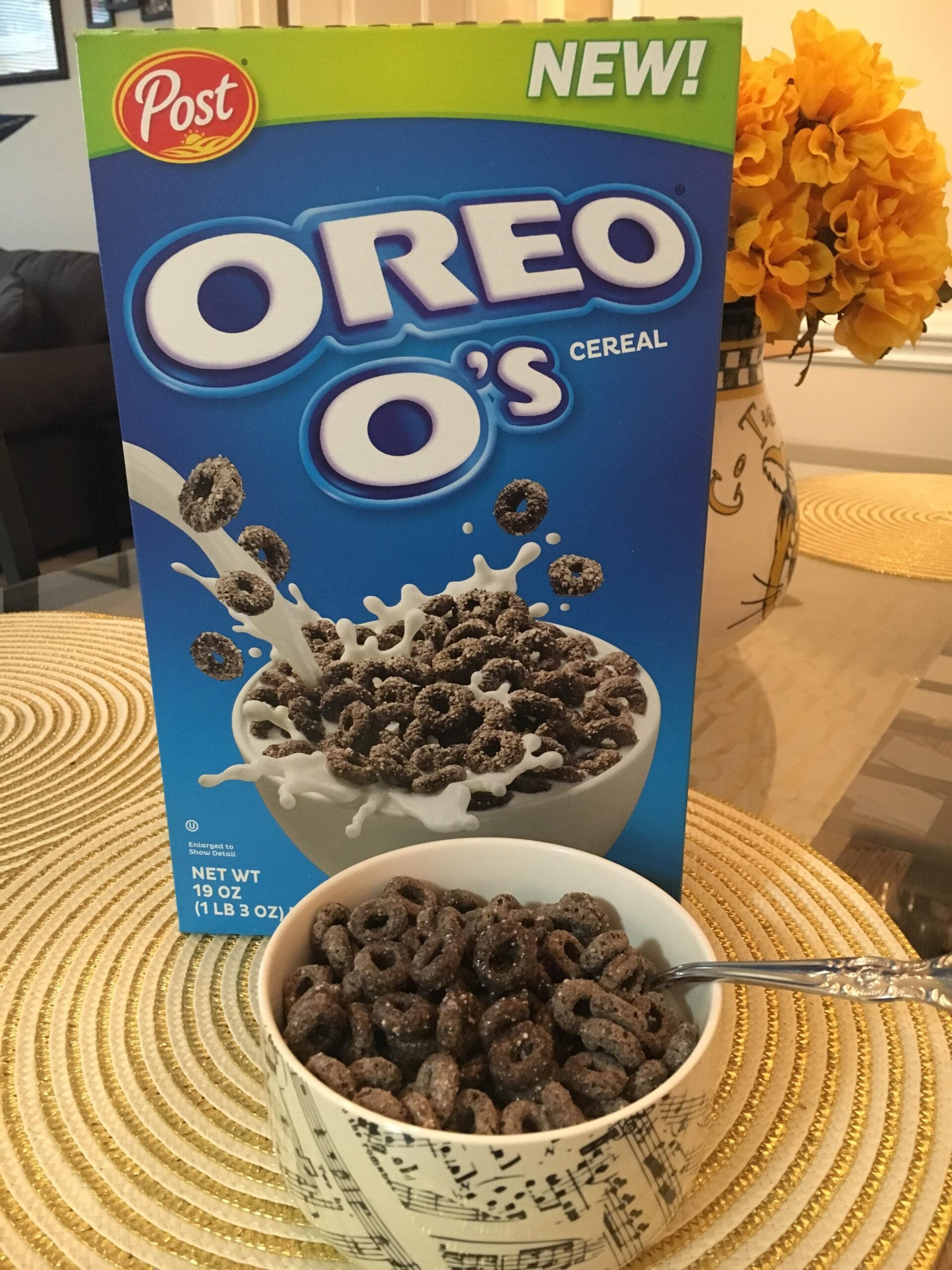 A box of Oreo O’s cereal stands behind a bowl of the cereal with milk on a round placemat. A spoon rests in the bowl, and a vase of yellow flowers is in the background.