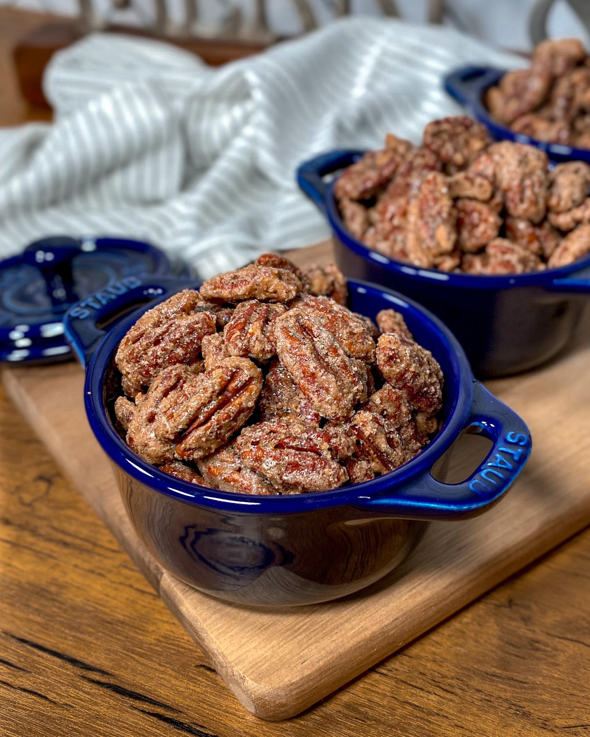 A blue ceramic bowl filled with candied pecans sits on a wooden board, with another similar bowl blurred in the background. A striped cloth is partially visible behind them.