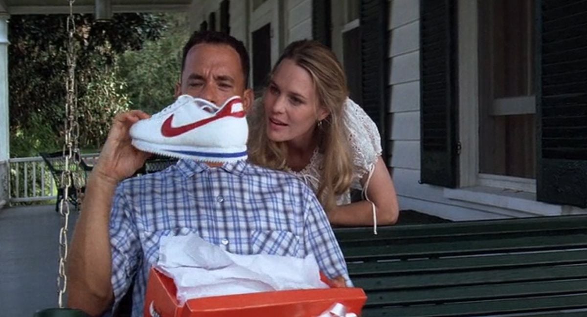 A man in a plaid shirt sits on a green porch swing, holding up a white Nike shoe with a red swoosh. A woman in white leans over his shoulder, looking at the shoe. Both appear to be examining the shoe from its box.