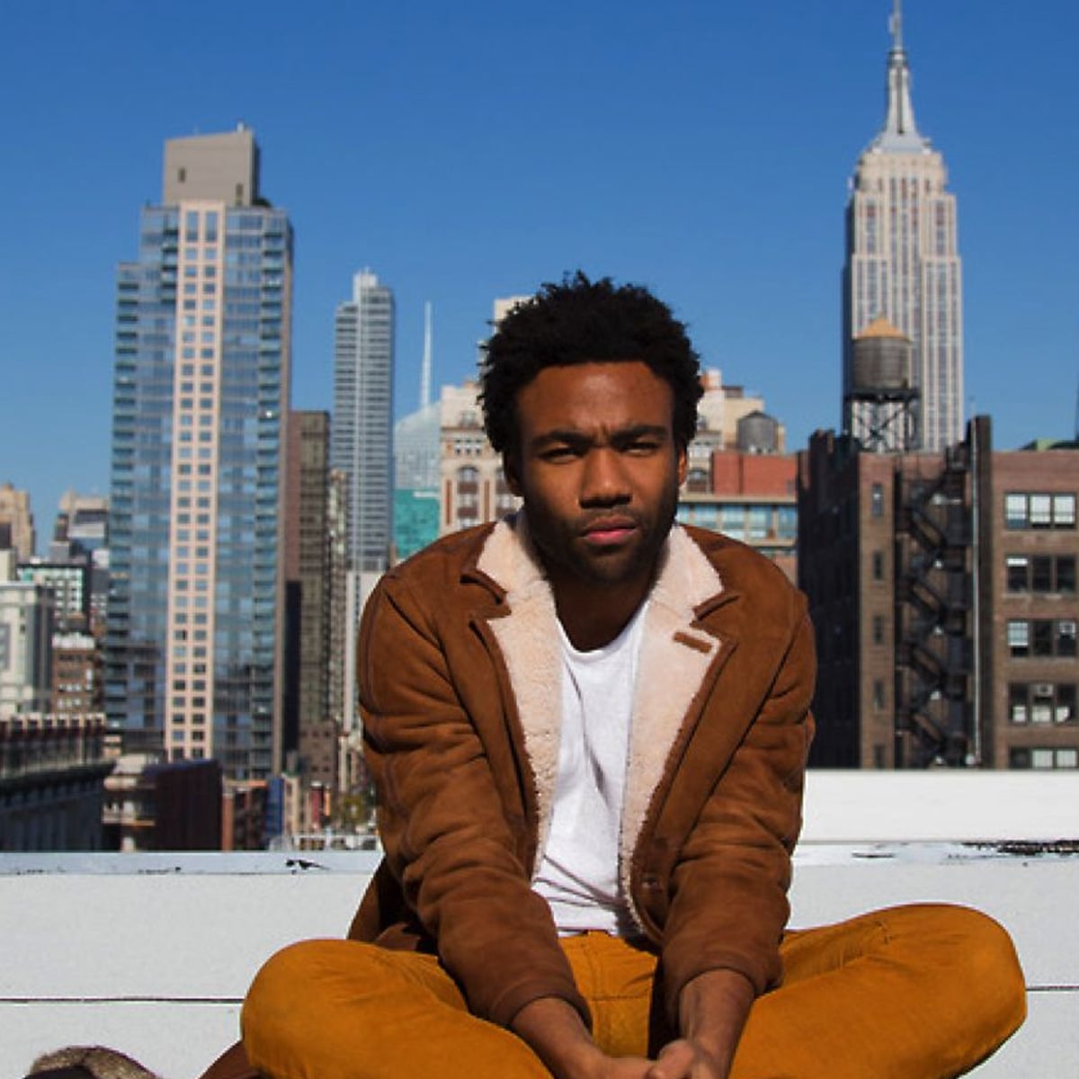 A man in a brown shearling jacket and orange pants sits cross-legged on a rooftop, with a clear blue sky and tall city skyscrapers, including the Empire State Building, in the background.