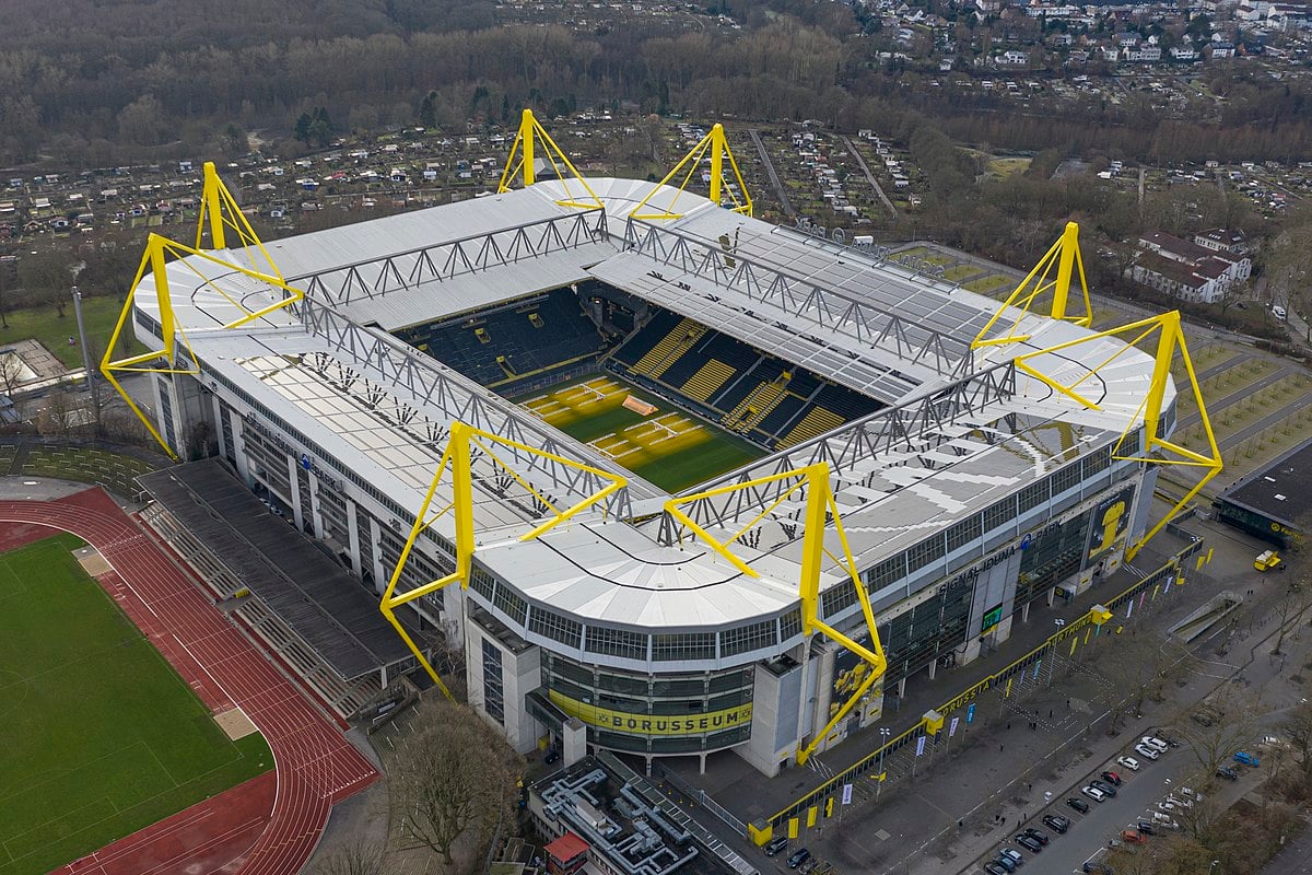 Aerial view of Signal Iduna Park, a large football stadium in Dortmund, Germany, featuring yellow steel supports and a green pitch inside, surrounded by stands and urban infrastructure.