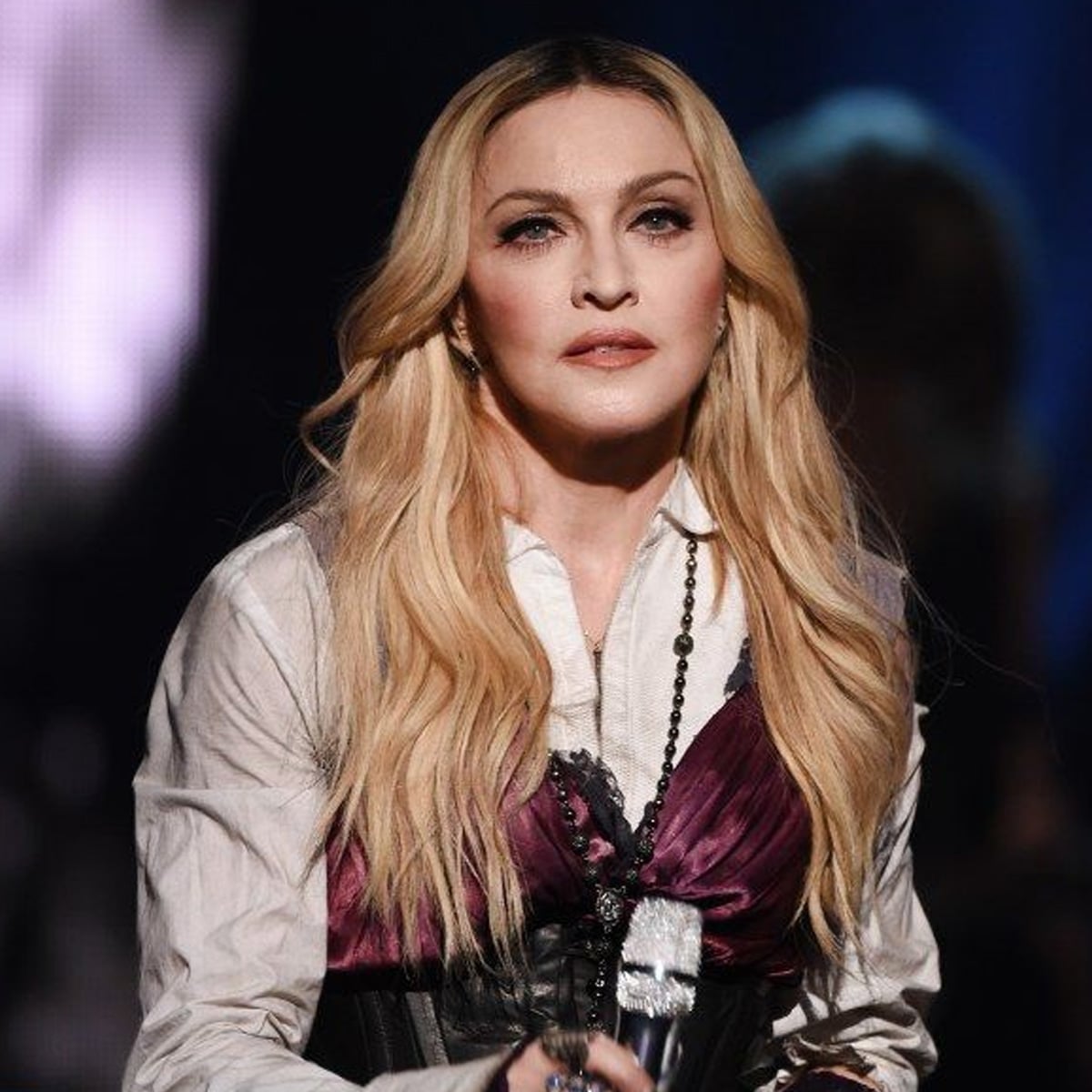 A woman with long blonde hair wearing a white shirt and a purple corset top holds a microphone on stage, with dark and blurred lighting in the background.