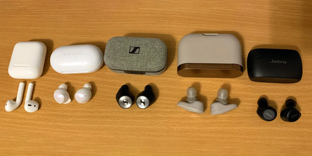 Six pairs of true wireless earbuds with their charging cases are neatly arranged on a wooden surface, with each pair of earbuds placed directly in front of its respective case.