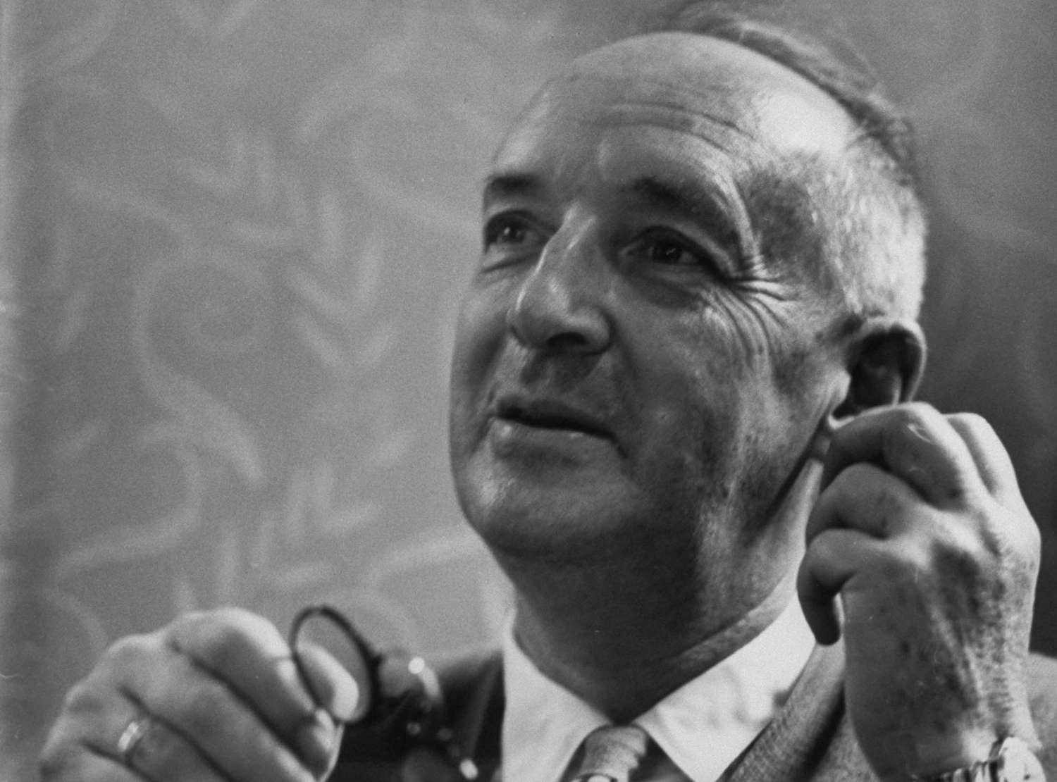 A middle-aged man with short hair looks upward thoughtfully, holding a pair of glasses in one hand and adjusting his ear with the other. He wears a suit jacket, shirt, and tie. The background is softly patterned.