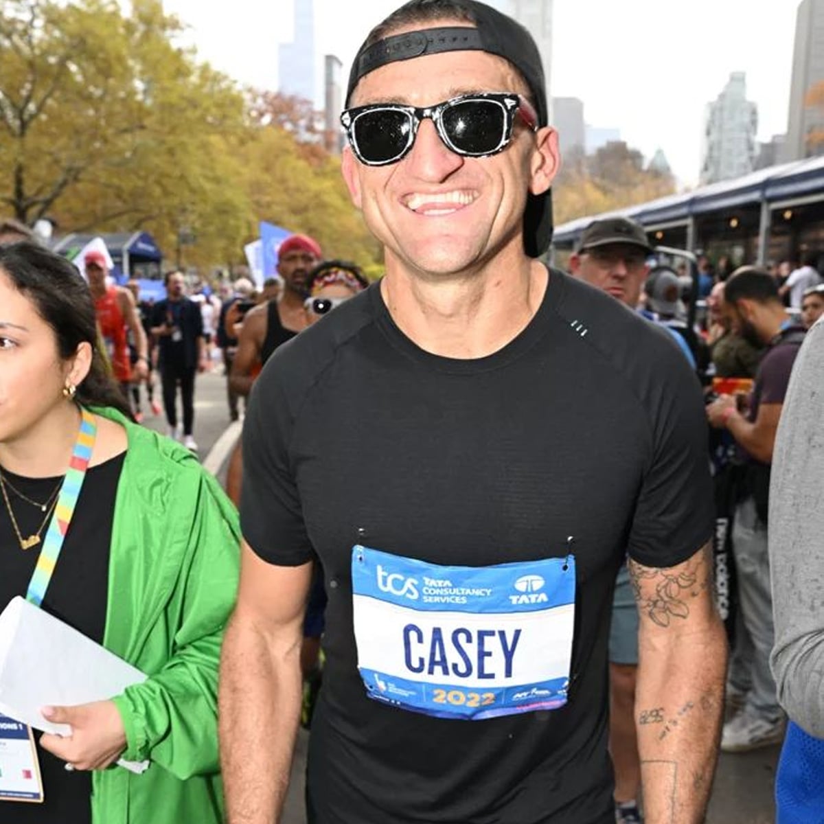 A smiling man wearing sunglasses and a black shirt with a “CASEY” race bib prepares to run in a marathon among a crowd of participants. Trees and city buildings are visible in the background.