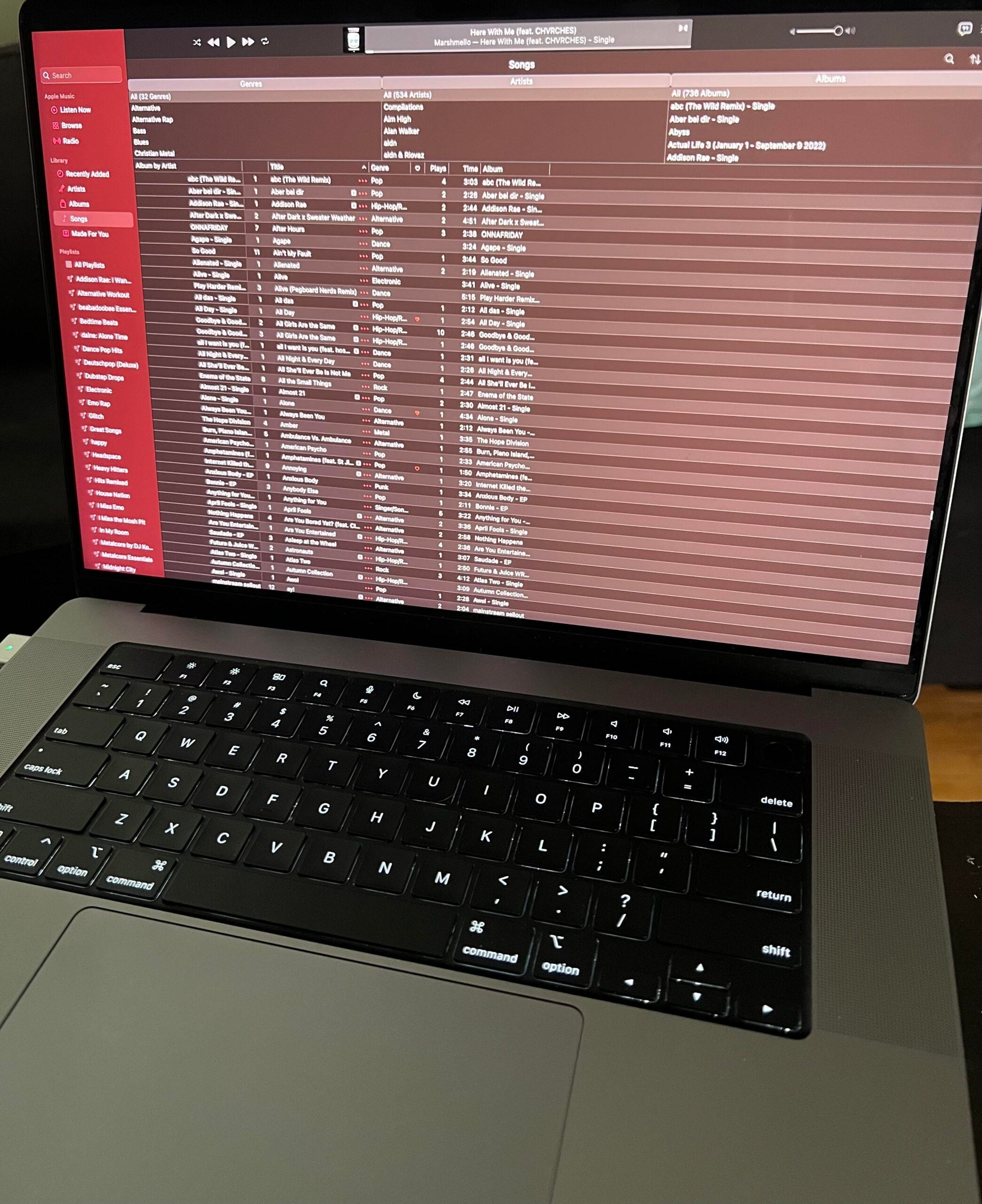 A laptop displaying a music library on its screen, with song titles, artists, and album information listed in a grid. The keyboard is visible in the foreground.