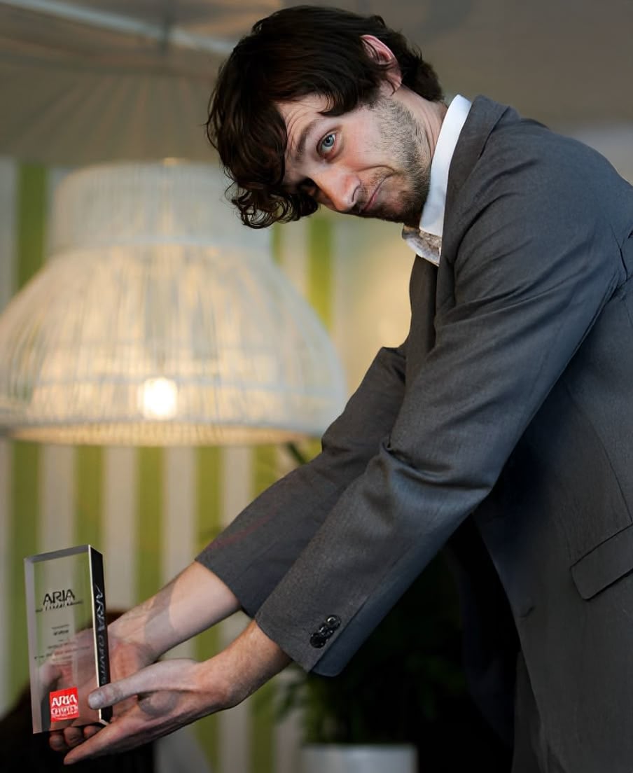 A man in a gray suit leans forward, smiling and holding an ARIA trophy with both hands. A large, white, woven lampshade and green-and-white striped wall are visible in the background.