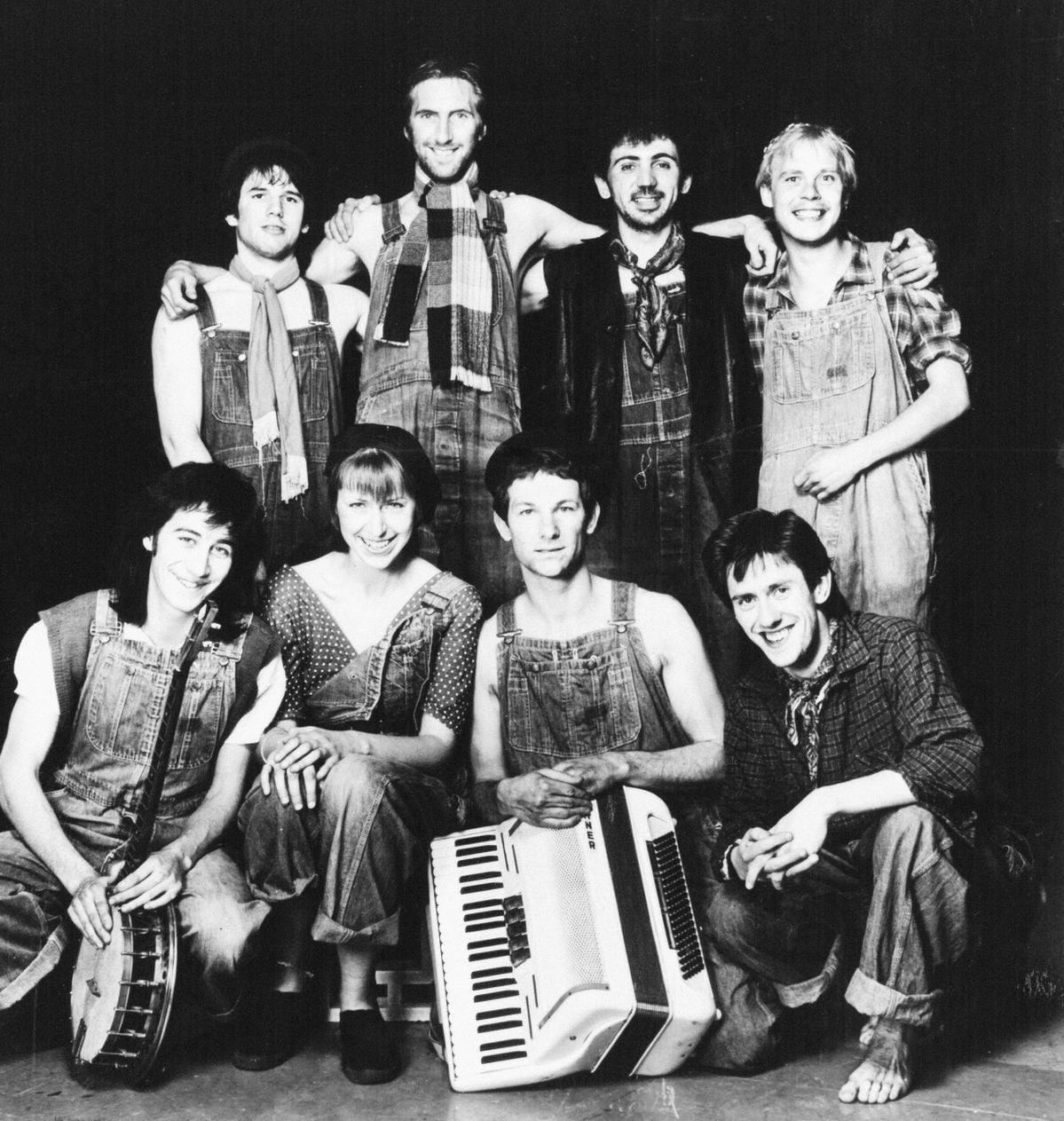 A group of eight people, wearing overalls and smiling, pose together. Some are standing while others are sitting, holding musical instruments including a banjo and an accordion. The photo is black and white.