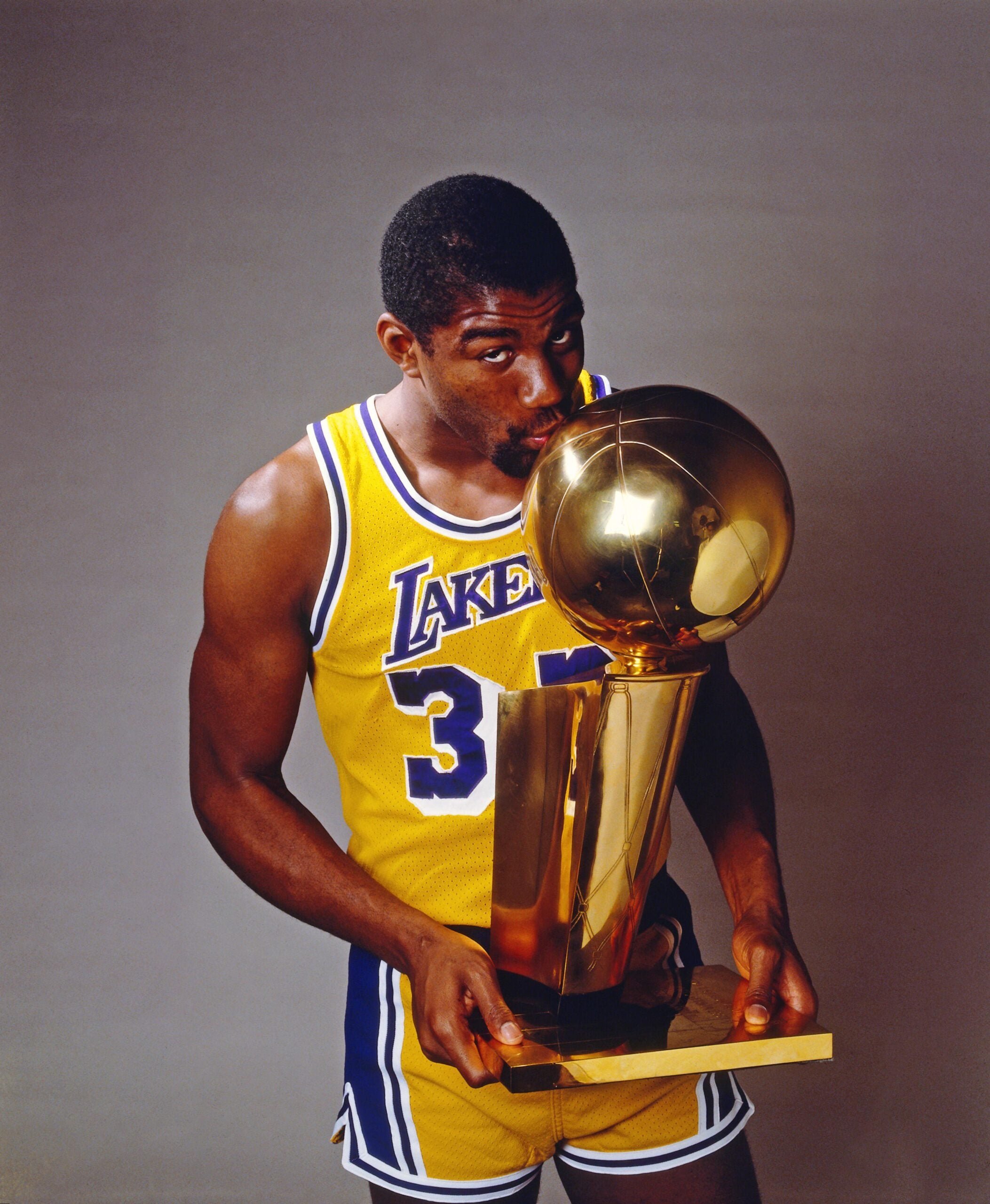 A basketball player in a yellow Lakers jersey with number 32 kisses a large gold trophy, celebrating a championship victory against a plain background.