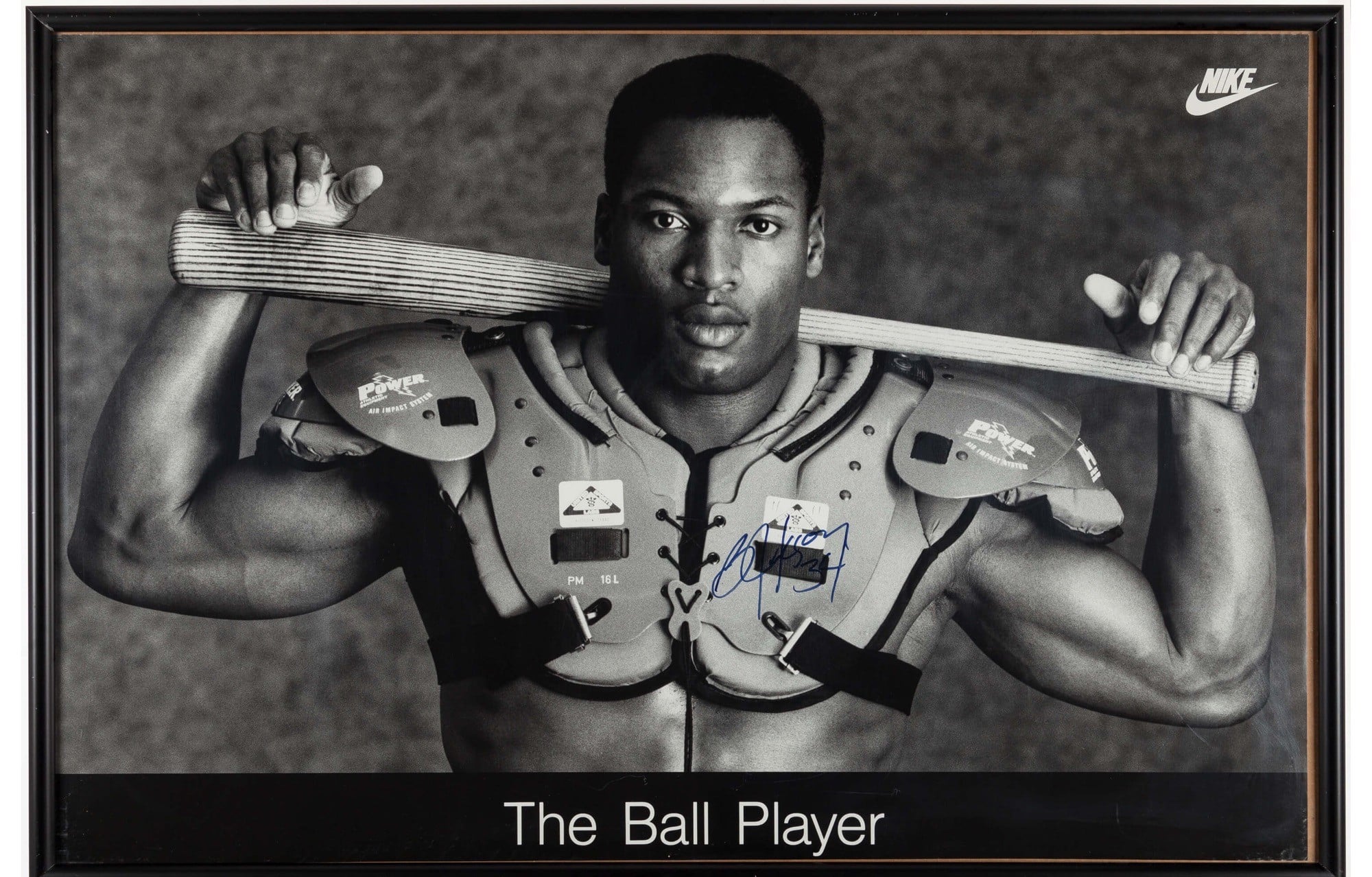 Black-and-white photo of a muscular man wearing football shoulder pads, holding a baseball bat across his shoulders, with a neutral expression. The image is labeled “The Ball Player” and features a Nike logo in the corner.