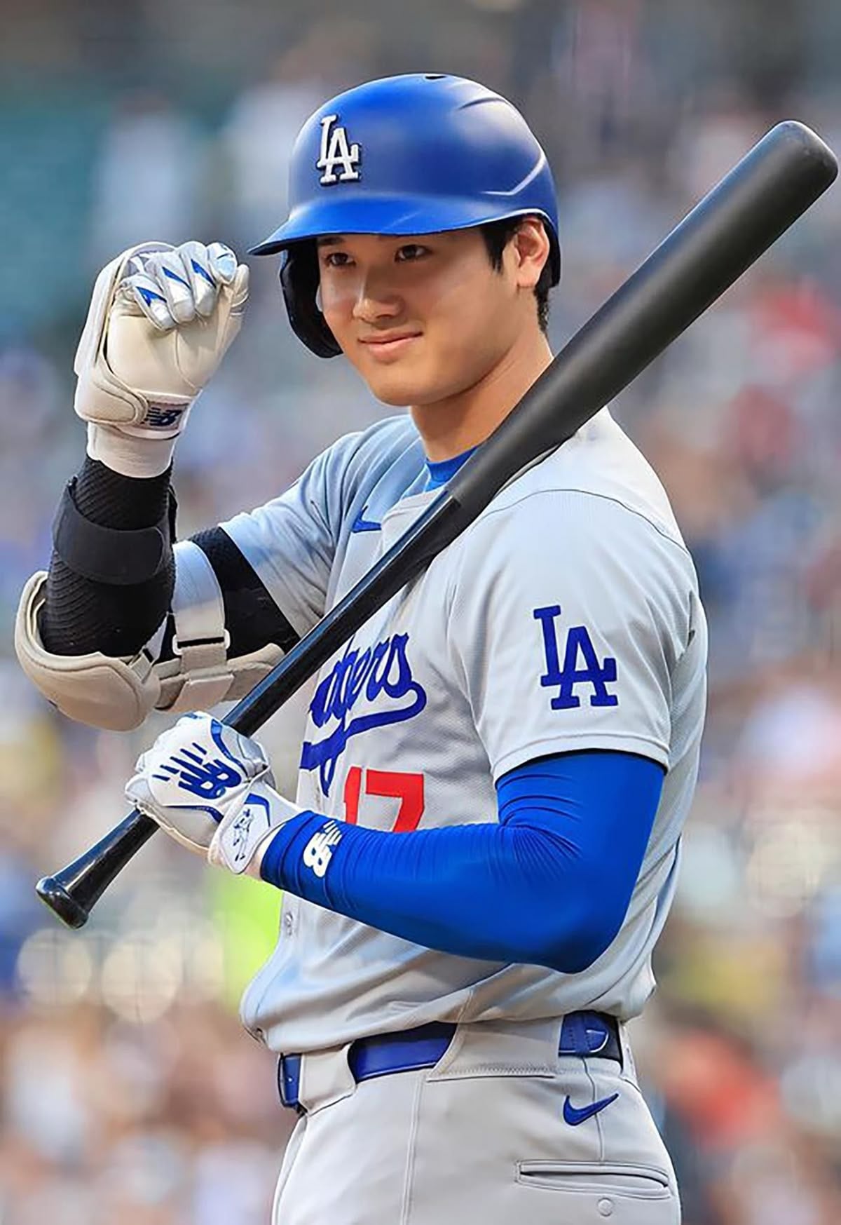 A baseball player in a Los Angeles Dodgers uniform and helmet smiles and gestures with his right hand while holding a bat over his left shoulder on a baseball field.