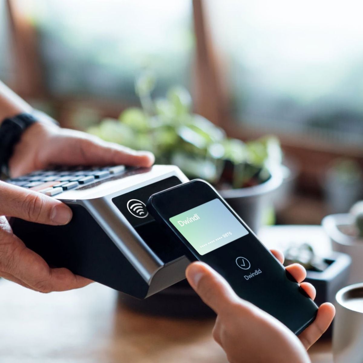 A person holds a smartphone near a card reader to make a contactless payment. The phone screen displays "Divided" and a progress bar, indicating the transaction is processing. Plants are blurred in the background.