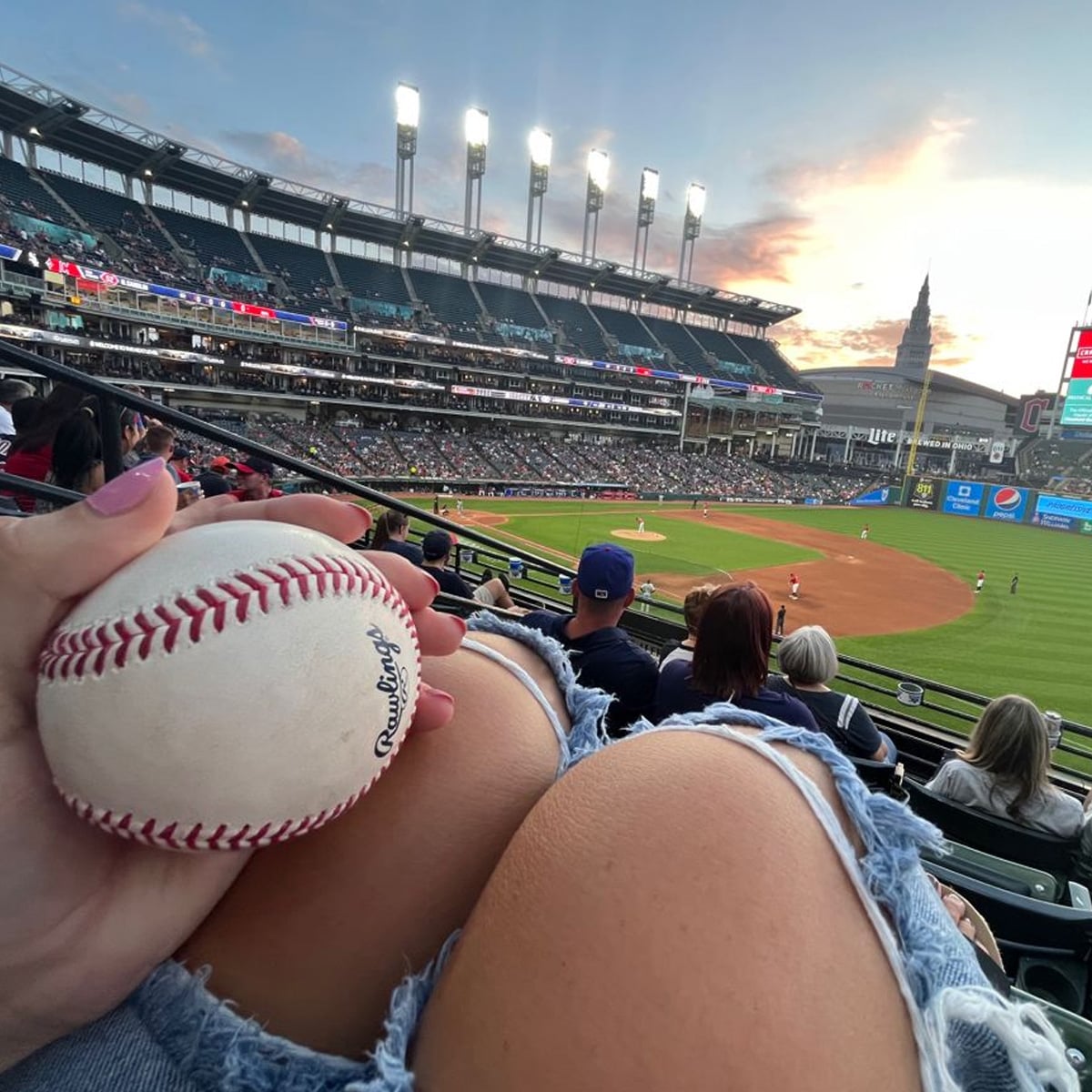 A person holding a baseball with chipped red nail polish, resting on ripped jeans at a stadium with a baseball game in progress and a sunset sky in the background.